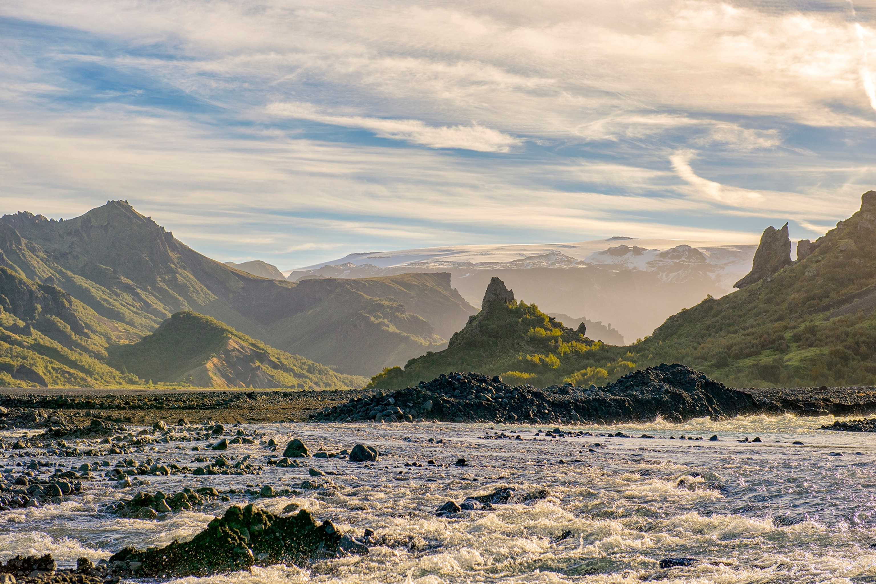 Fast flowing river backdropped by lush green mountains and a sunlit partly cloudy sky