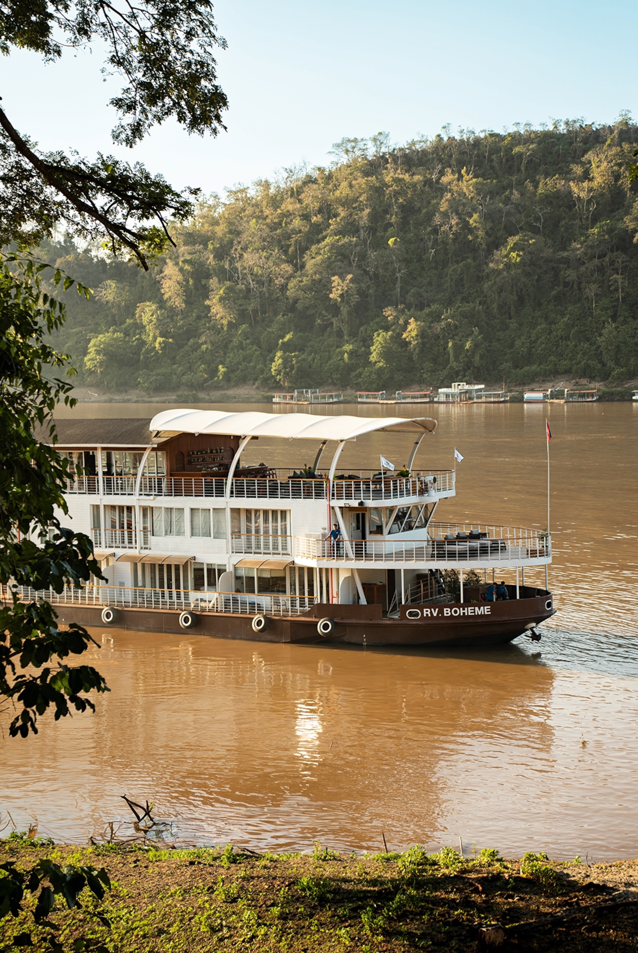 A three-decked river cruise boat going down a wide river with jungle on either side.