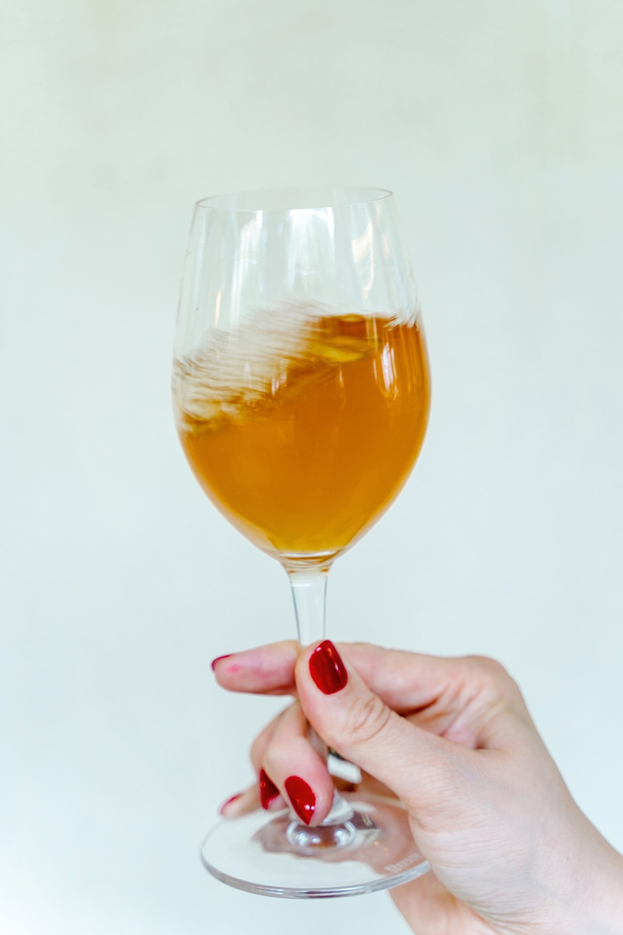 A hand with manicured nails holding up a glass of unfiltered orange wine against a plain wall.