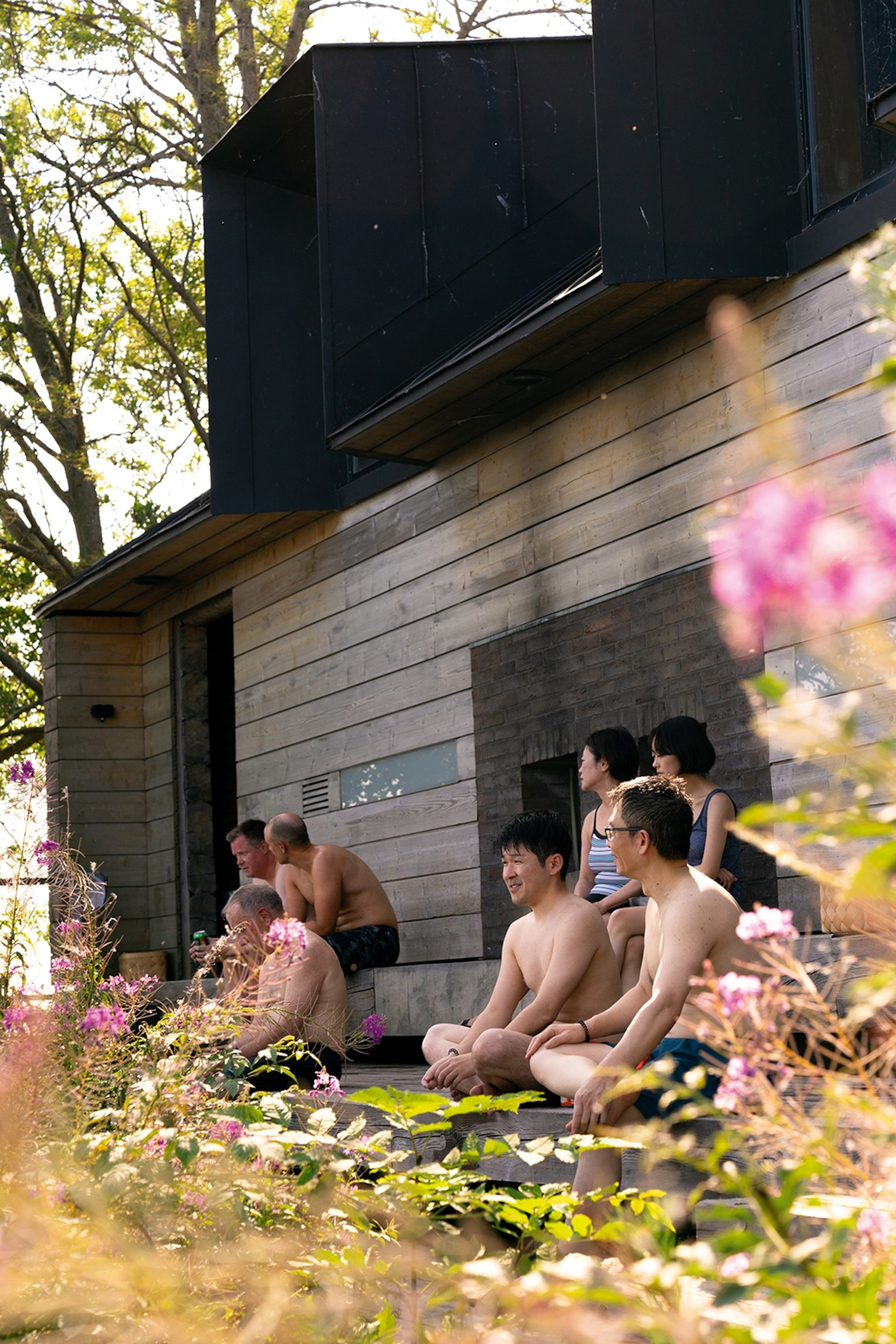 A wide range of people resting outside a wooden sauna, shot through flowering bushes as they gaze out towards the hidden lake ahead.