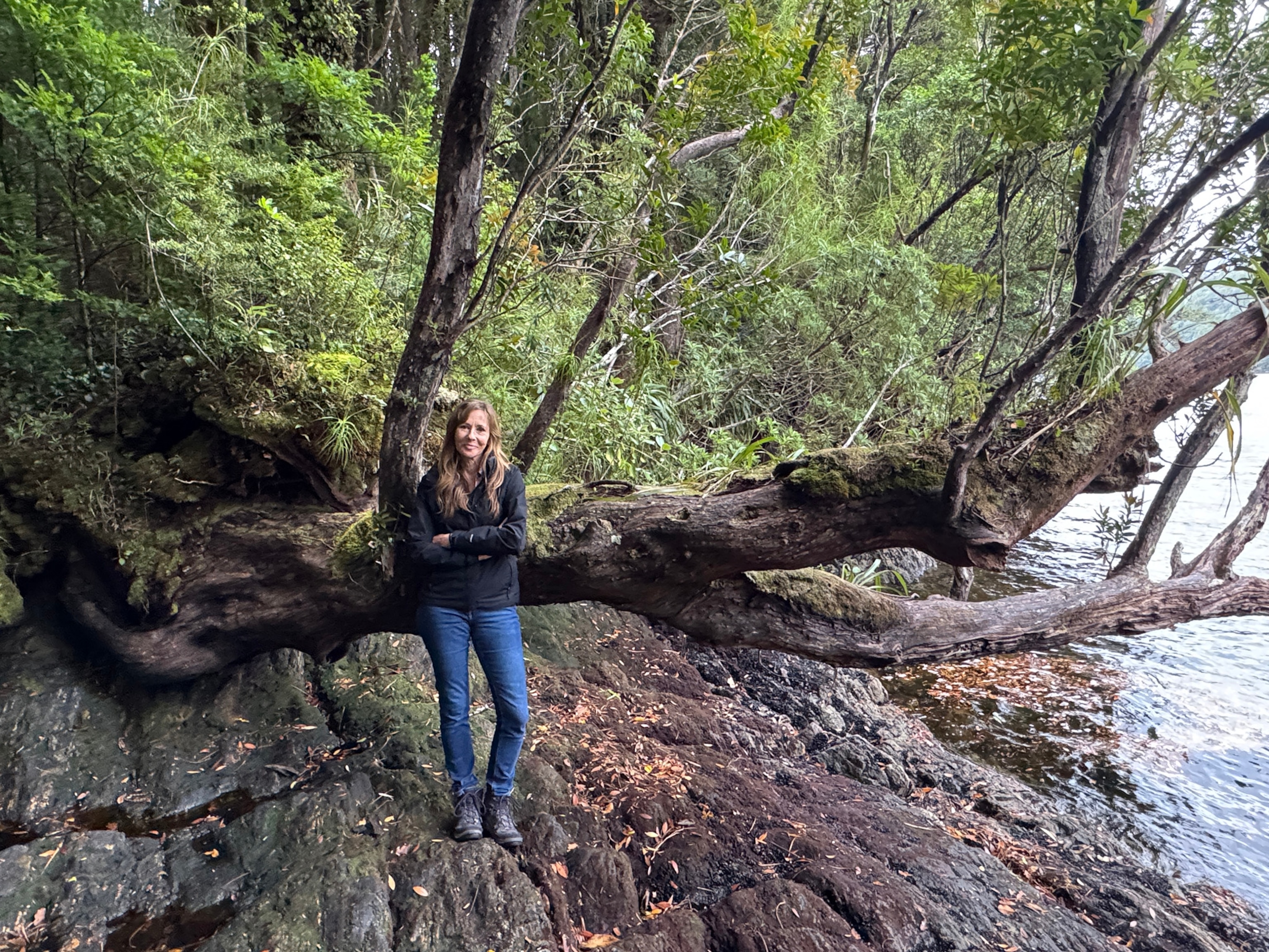 A woman wearing blue jeans and a black rain jacket leans against a tree, smiling at the camera.