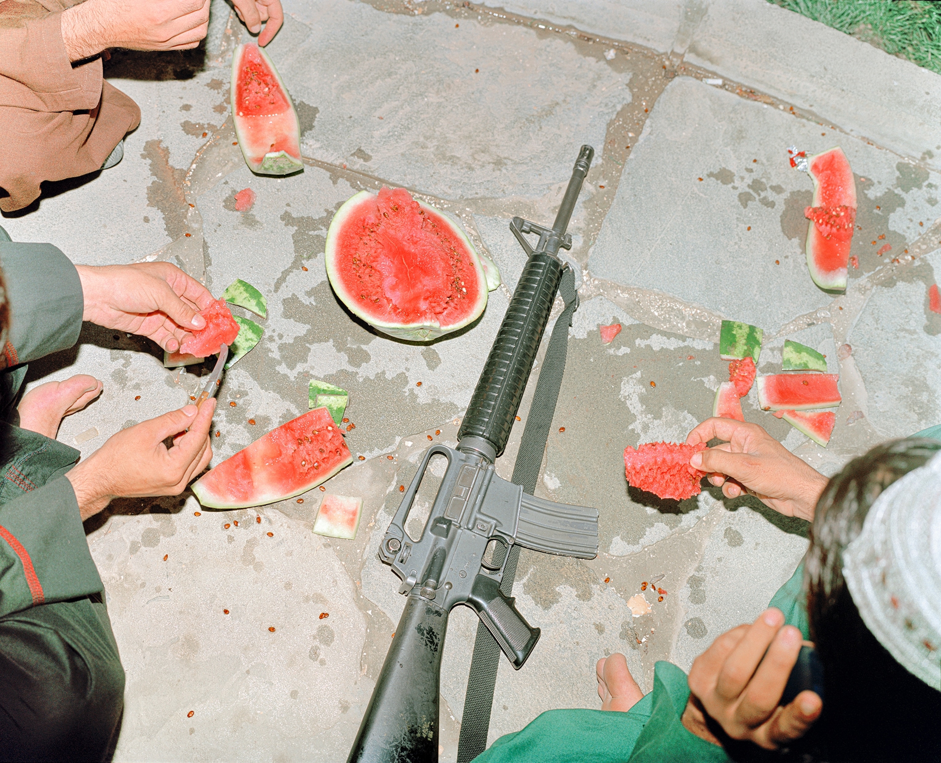 The hands of two Taliban fighters reach for sliced watermelon, while a gun sits on the ground in the middle of the pair.