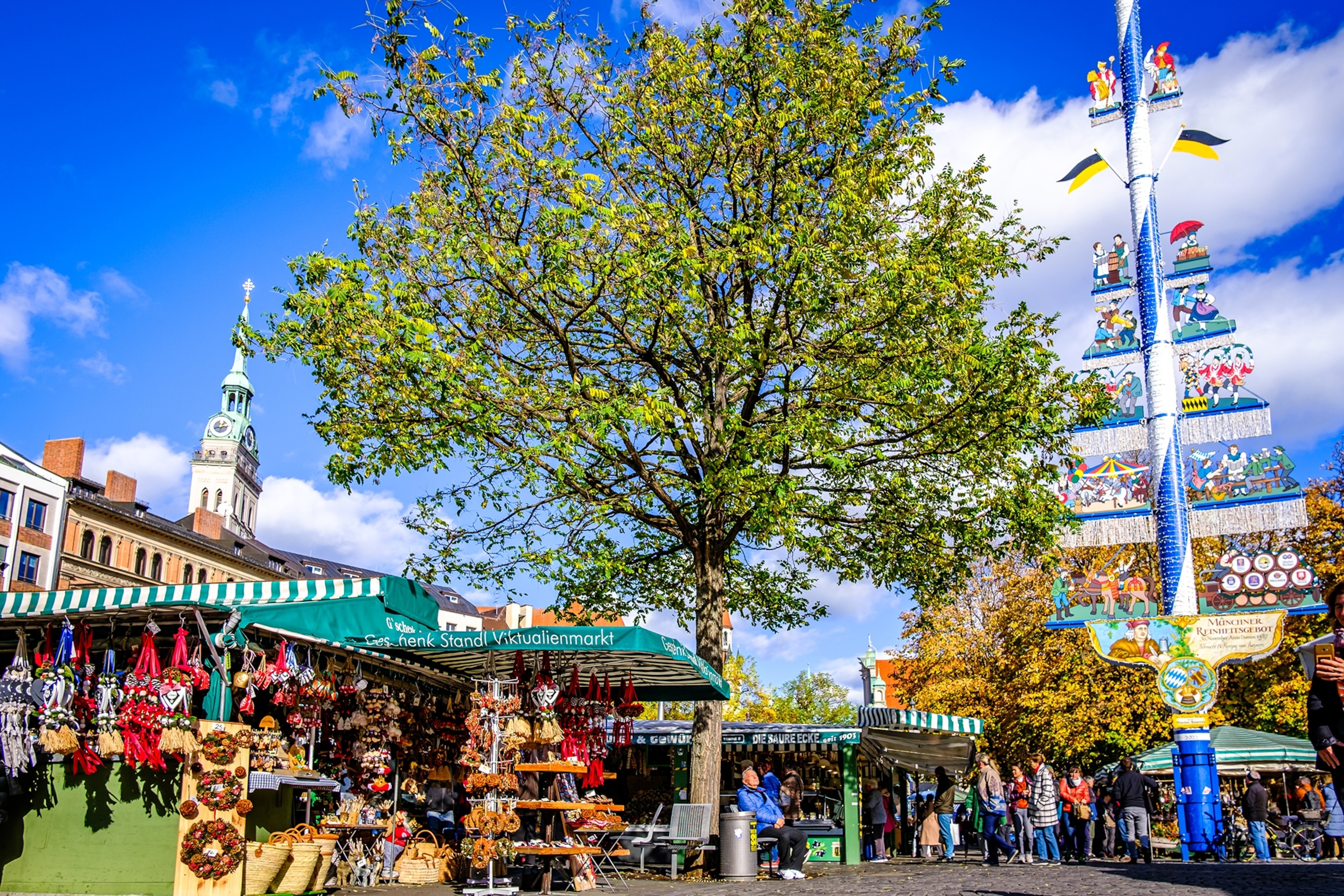 A sunny shot of a busy street and produce market, with a maypole and lush tree in the foreground.