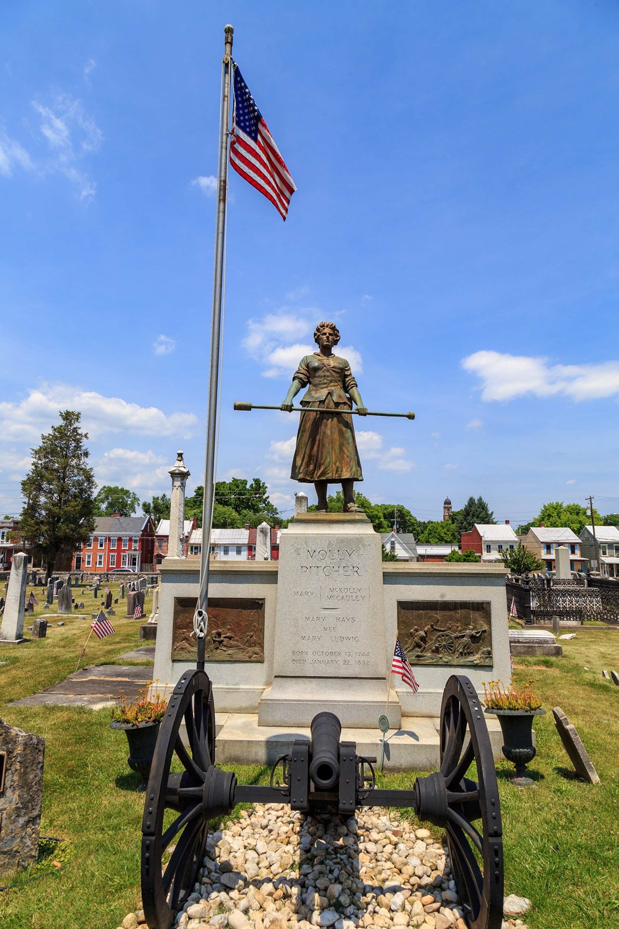 A bronze statue of a woman holding a long staff stands atop a plinth in a cemetery, an old canon in front and a tall flagpole waving an American flag behind