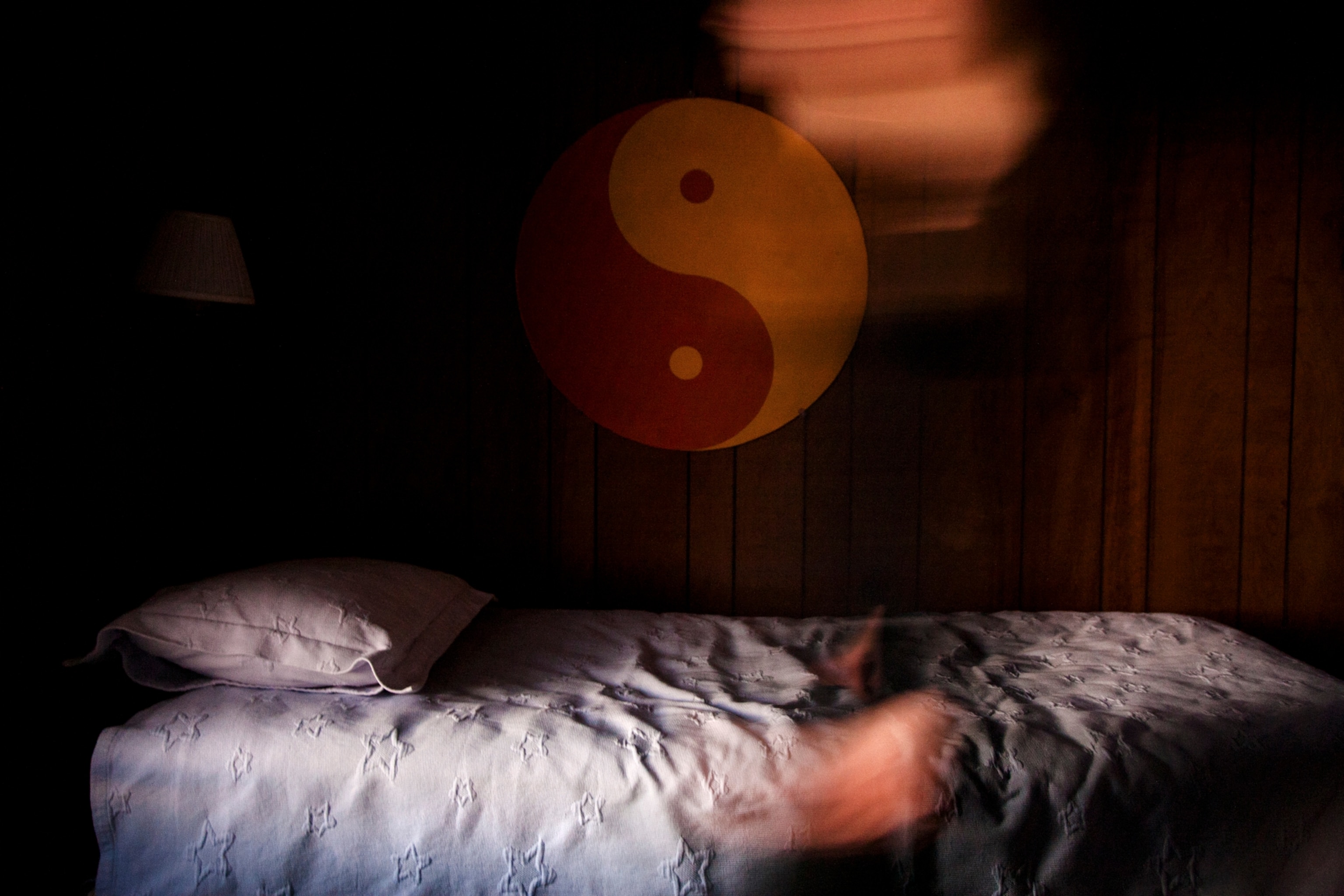 an orange and yellow yin yang hanging on the wooden wall over a white bed, golden window light falling on the wall and the bed