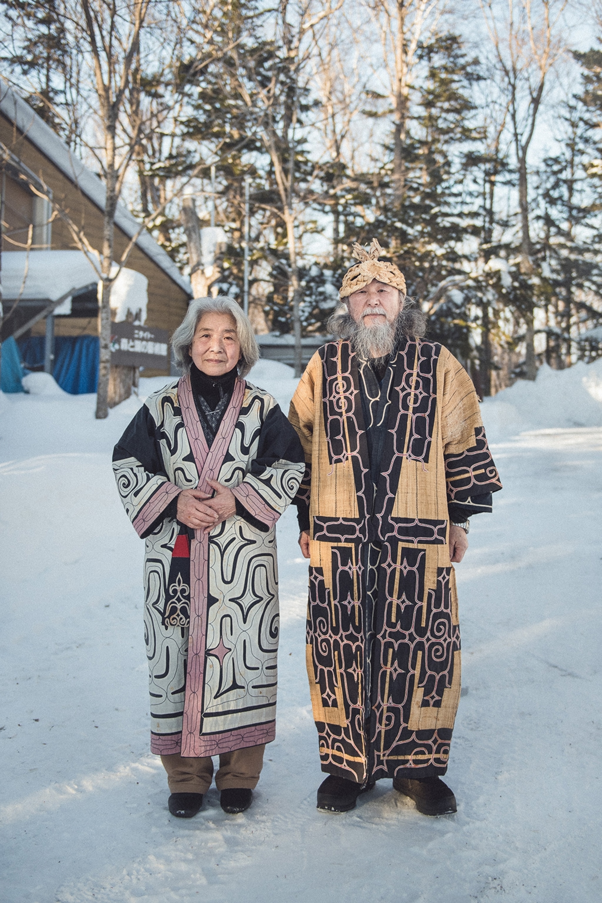 An elderly, smiling couple in traditionally embroidered yet casual kimonos, standing in the snowy landscape of a small village.