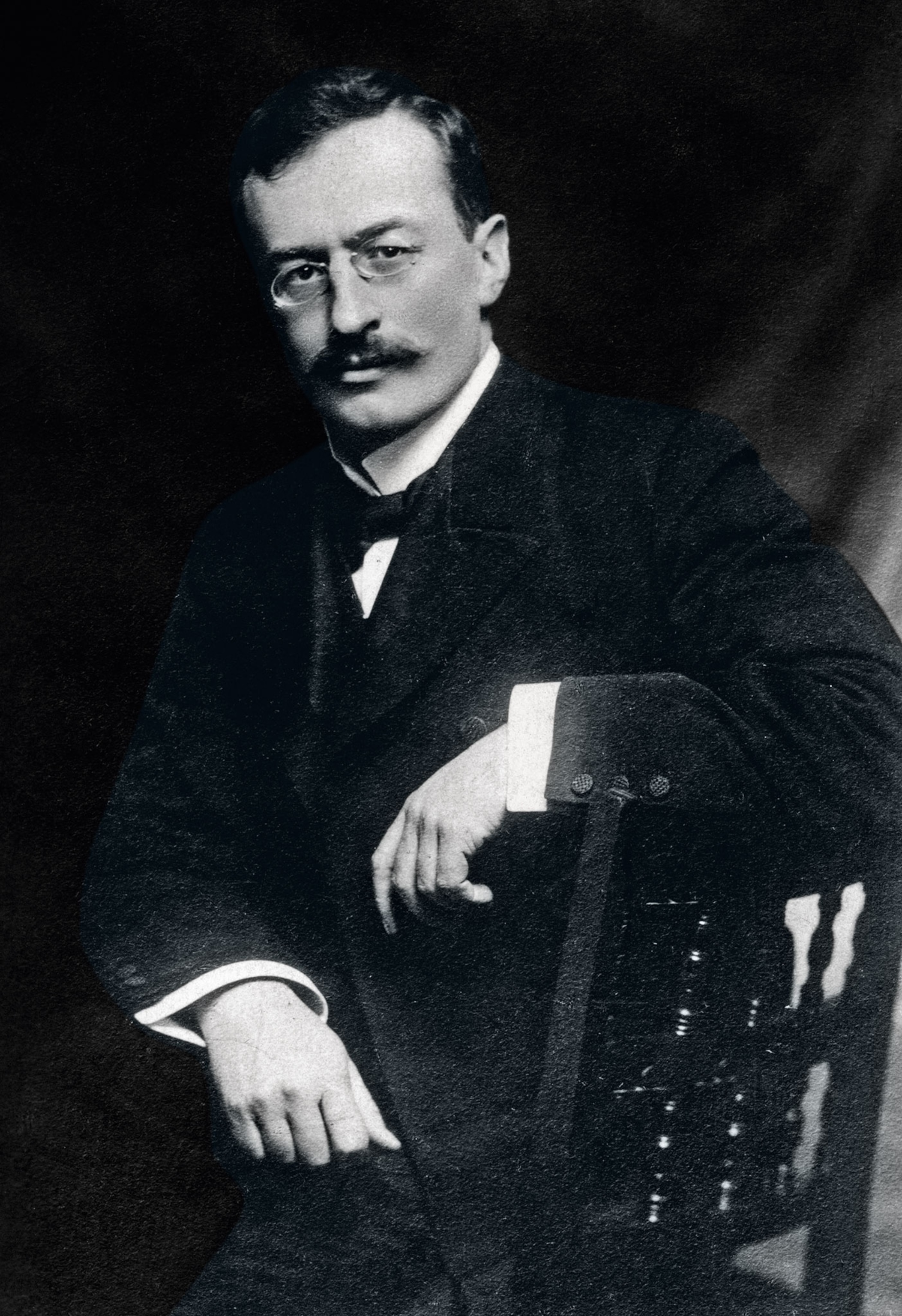 A man wearing glasses with a mustache poses in a suit in a black and white photo