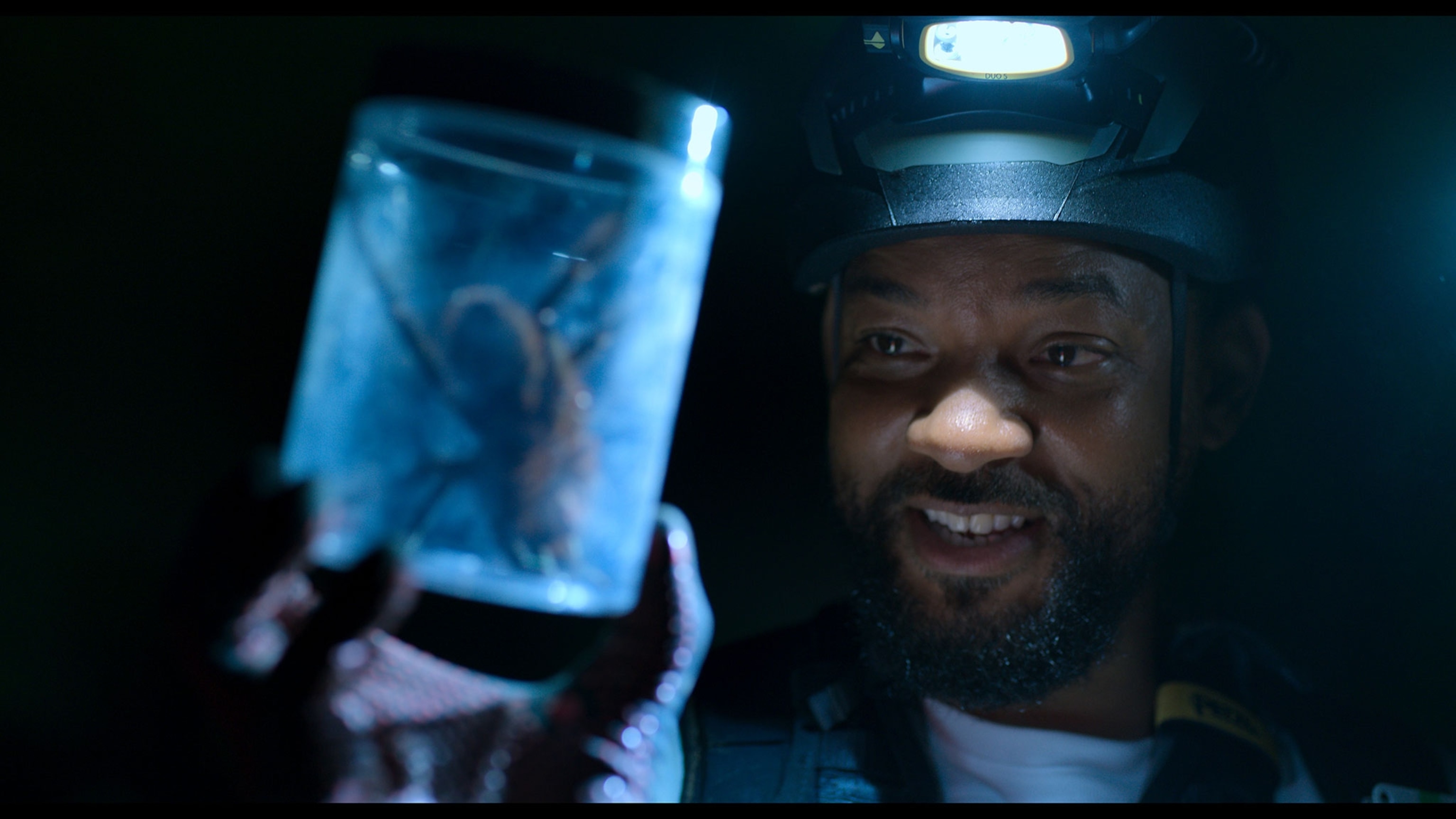 A man with dark skin has a headlight and holds a jar with a tarantula.