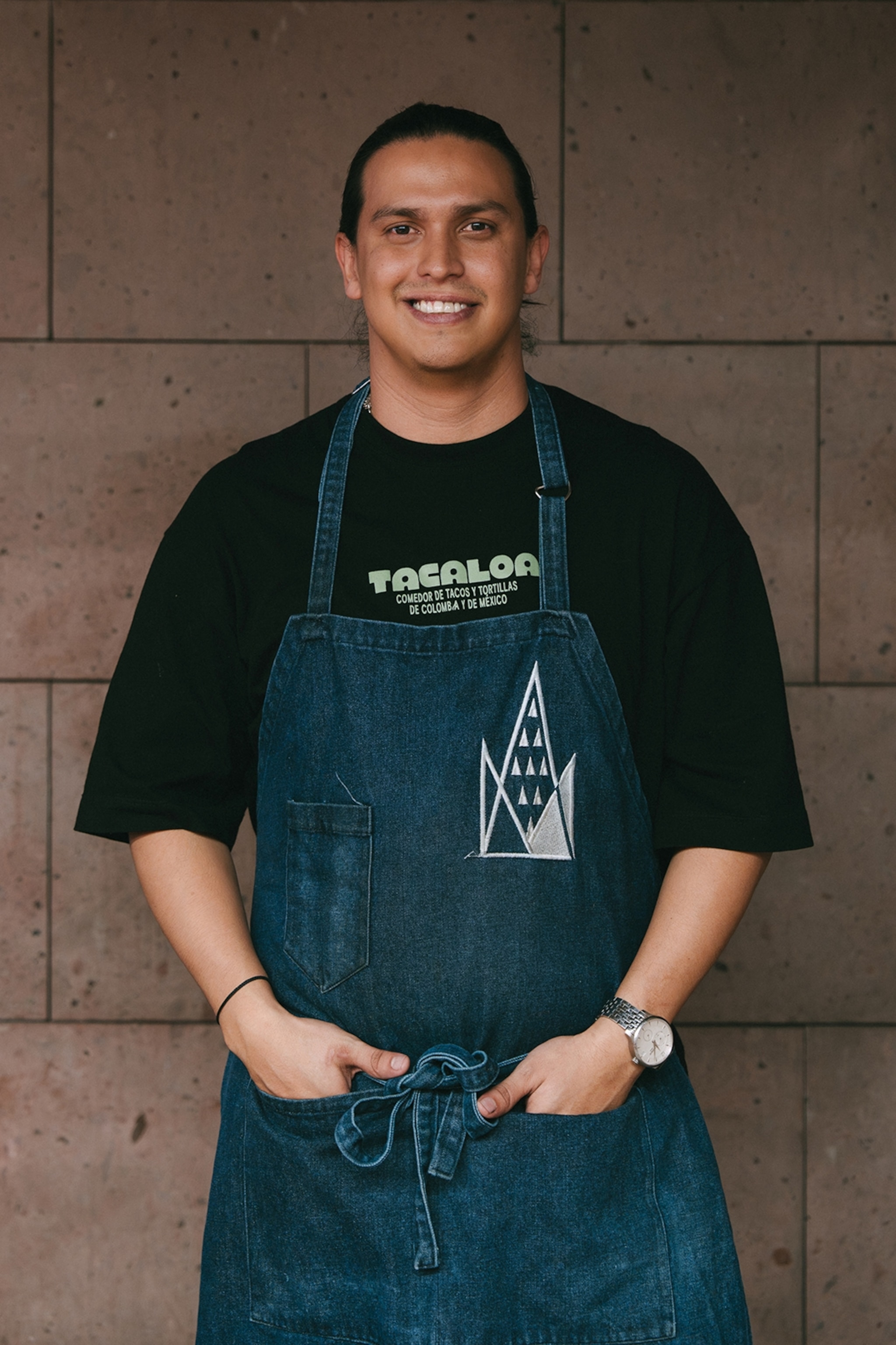 A smiling, young male chef wearing an apron and his long hair tied back into a bun.