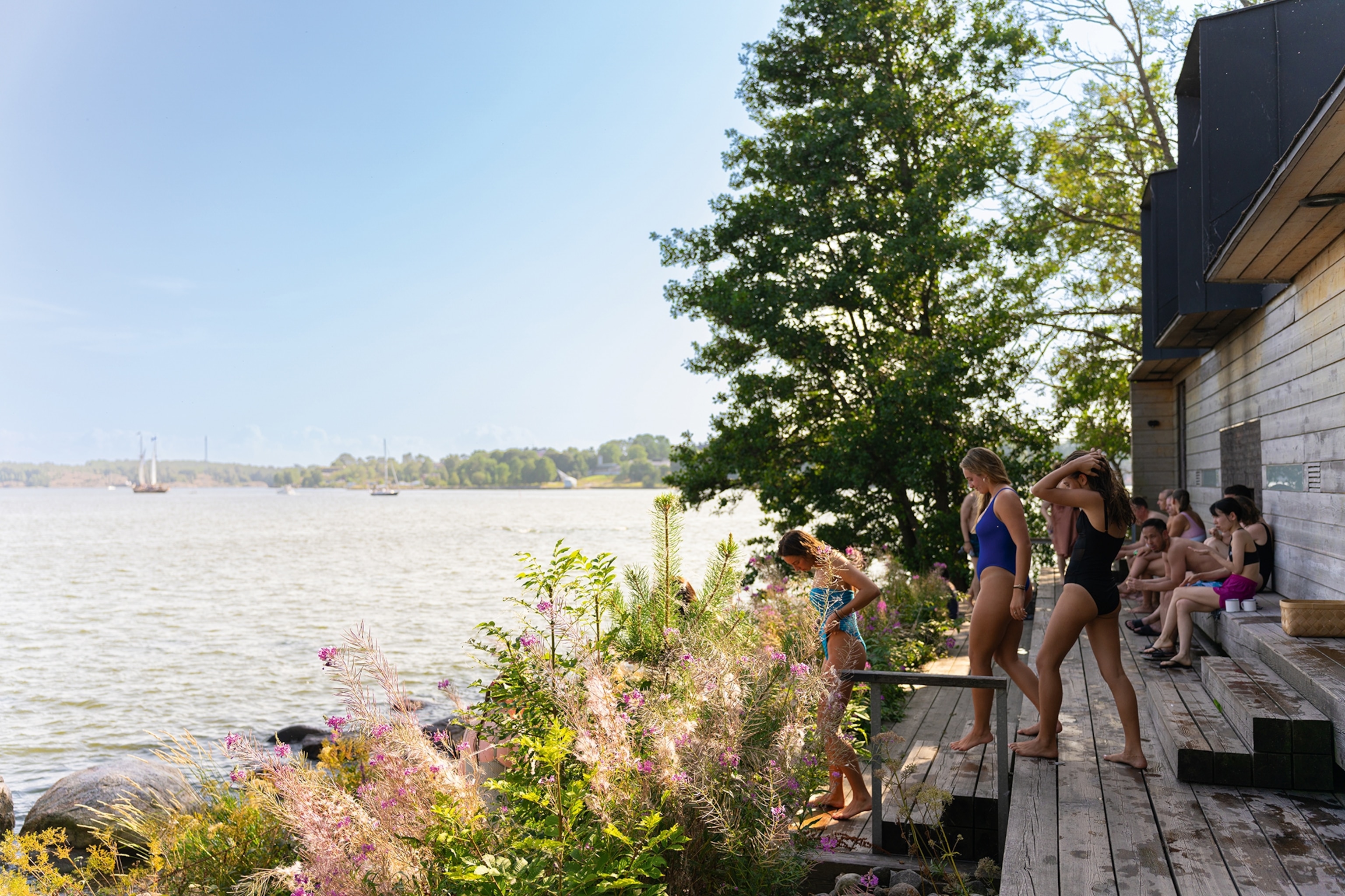 A dynamic landscape shot of lake-side sauna with a wooden terrace and pier leading into the water, surrounded by flower bushes.