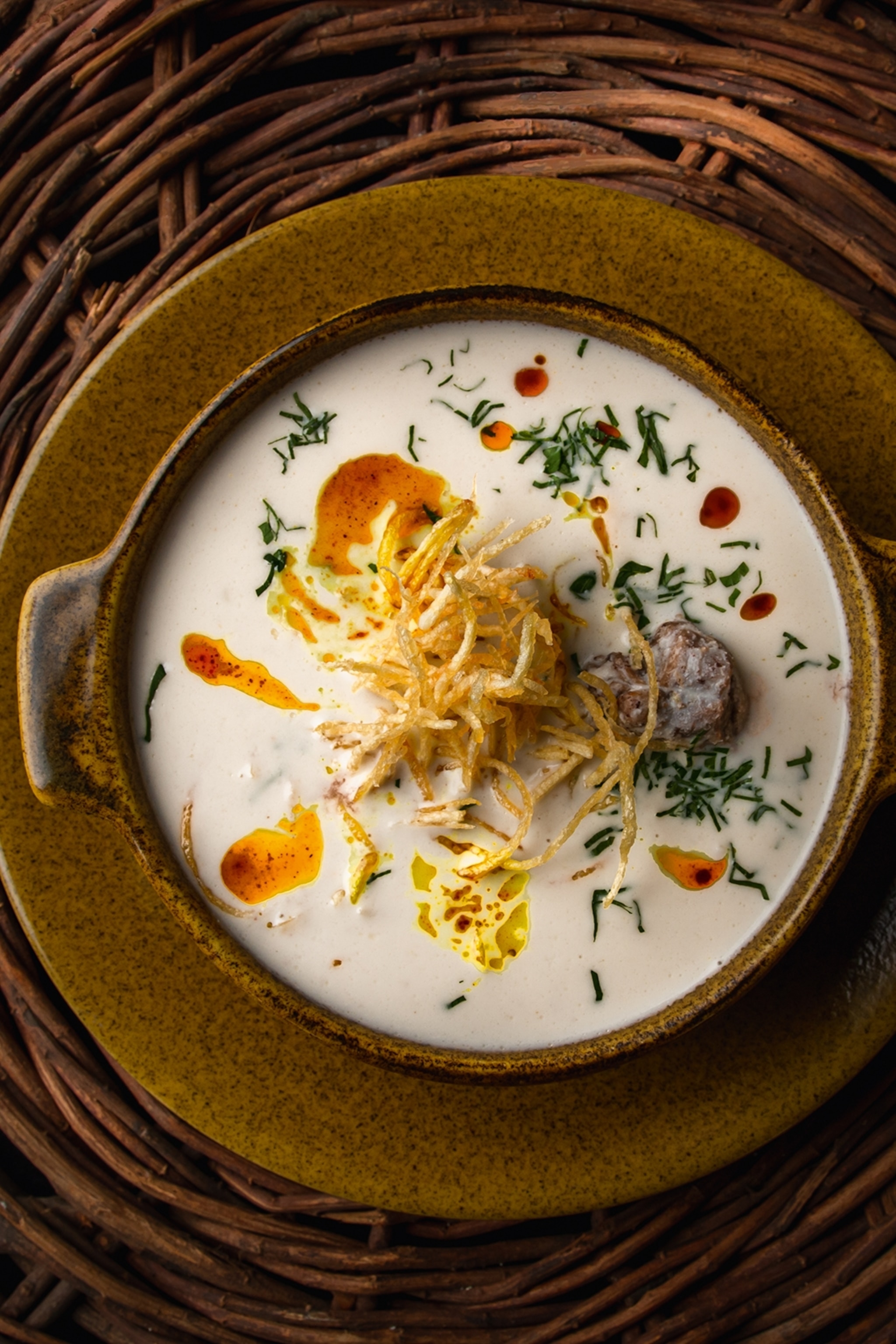 A fancy bowl of milky broth with crispy fried potato strips, herbs and a spice oil drizzled on top.
