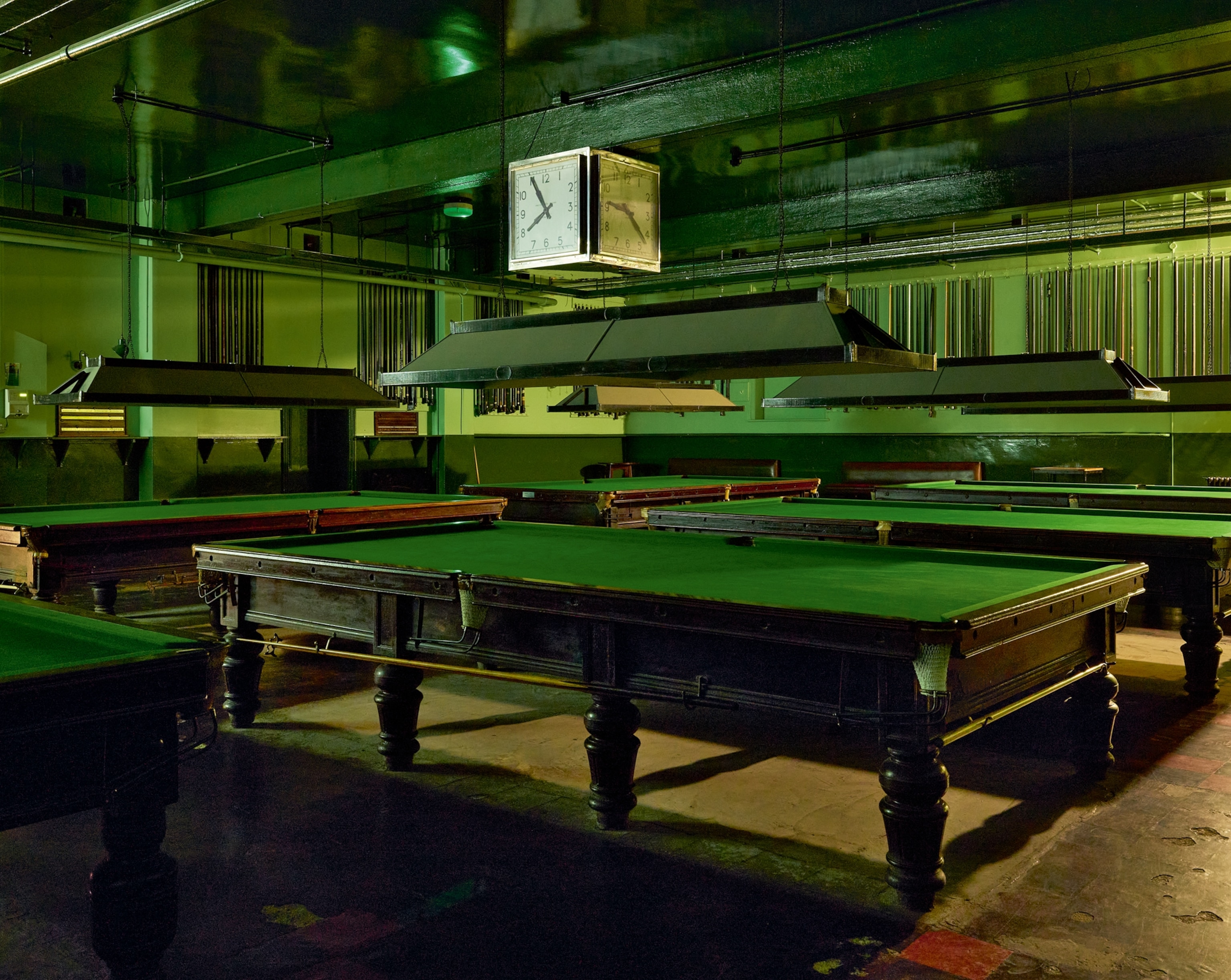 Dimly lit billiard hall with vintage green-felt tables and low-hanging lights. A square clock is prominently displayed on the ceiling, evoking a nostalgic mood.