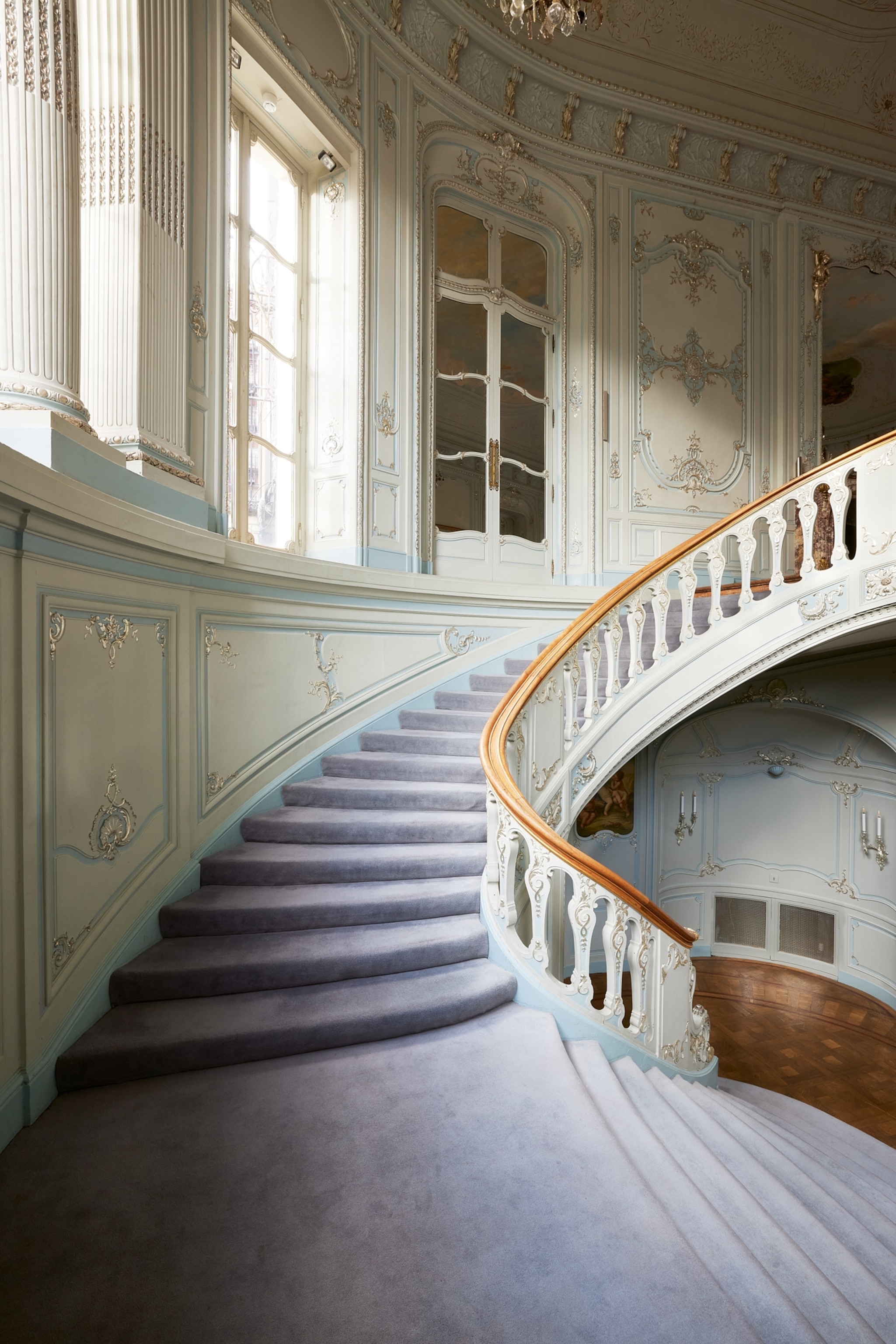 Elegant spiral staircase in a lavish interior, with ornate white walls and gold accents. Soft purple carpet adds a touch of warmth and luxury.