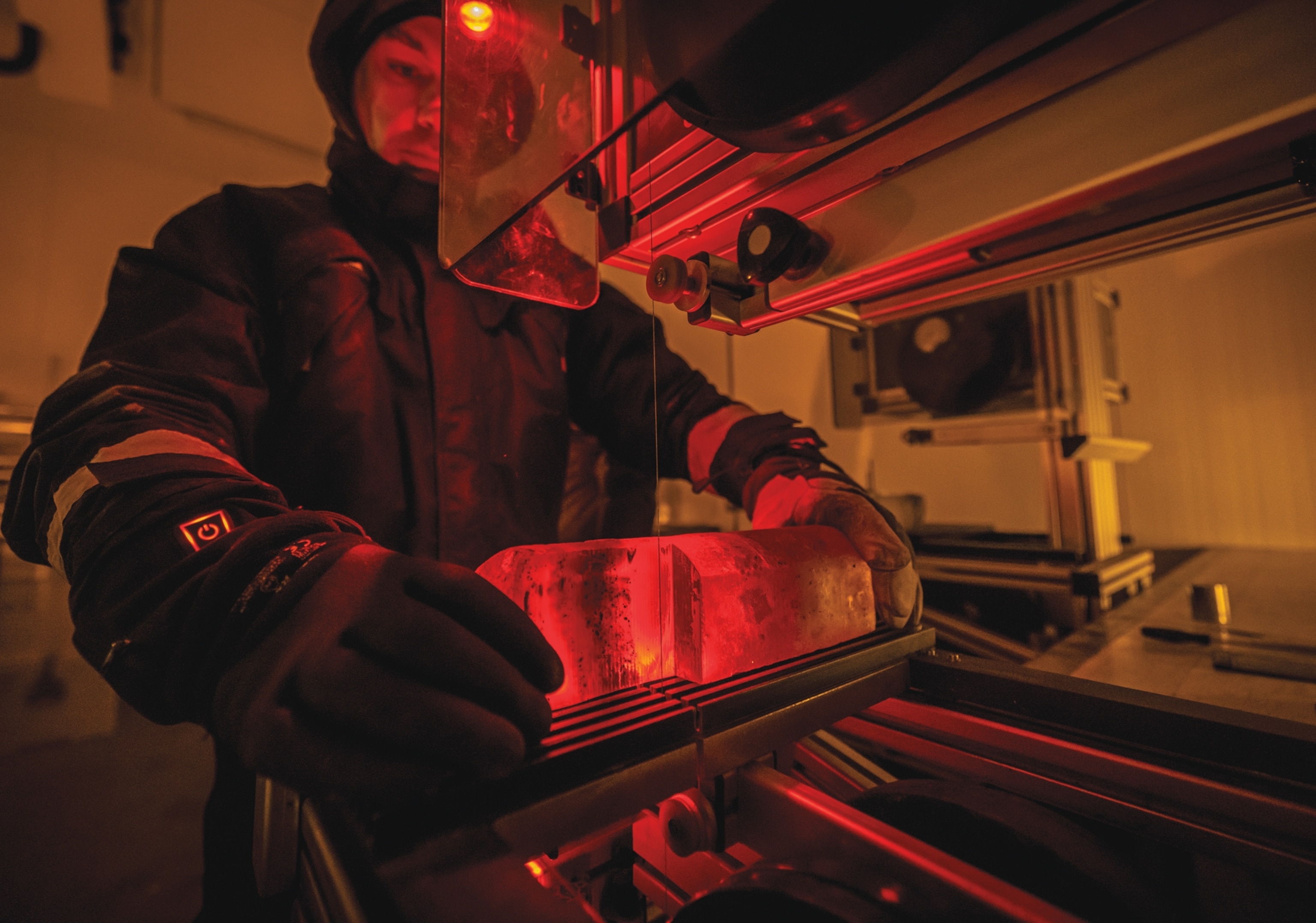 Person dressed in heavy coat and gloves holding a cylindrical block of ice under red light.