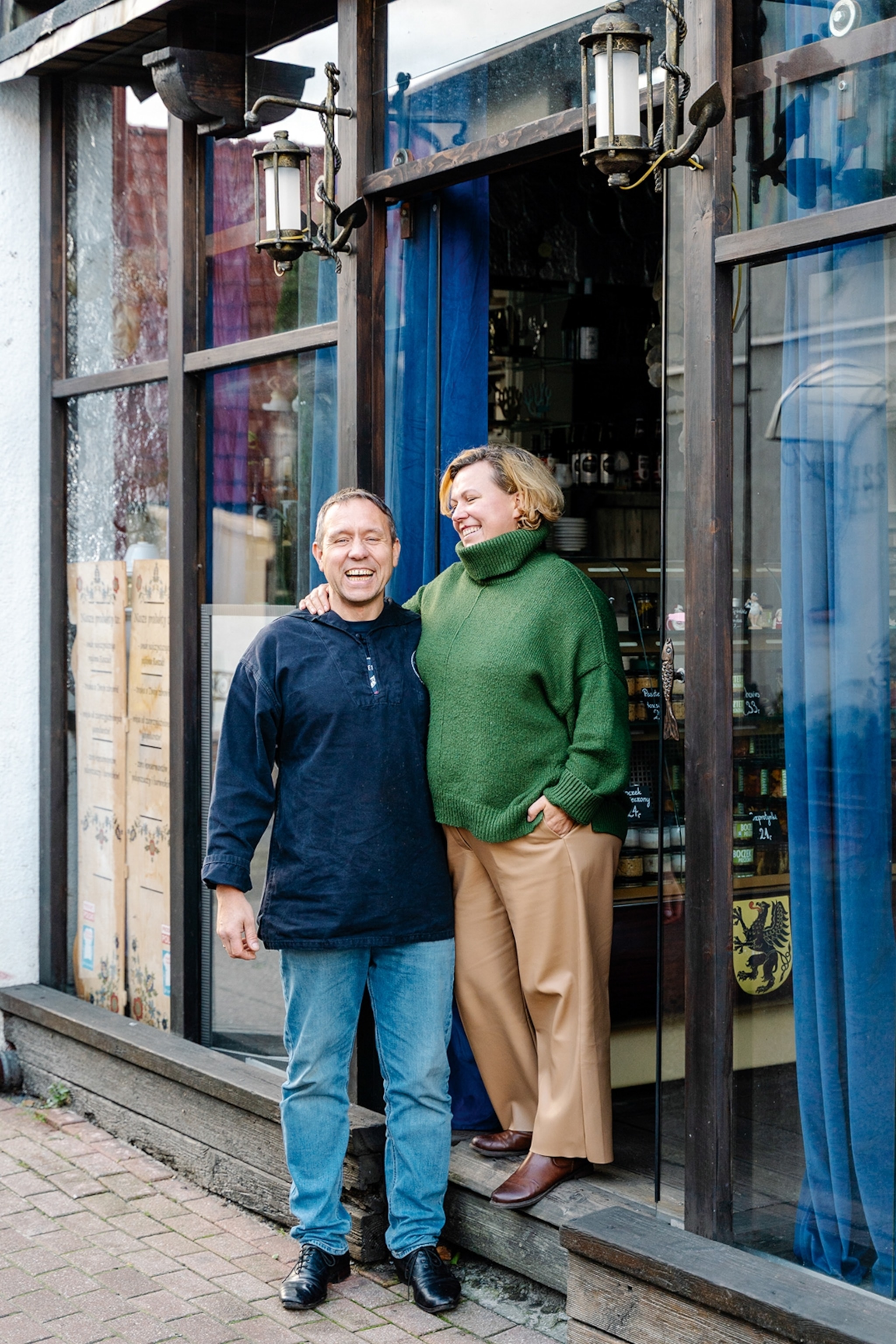 A local couple smiling into the camera, standing in the door of their street-side restaurant with wooden-framed entrance.