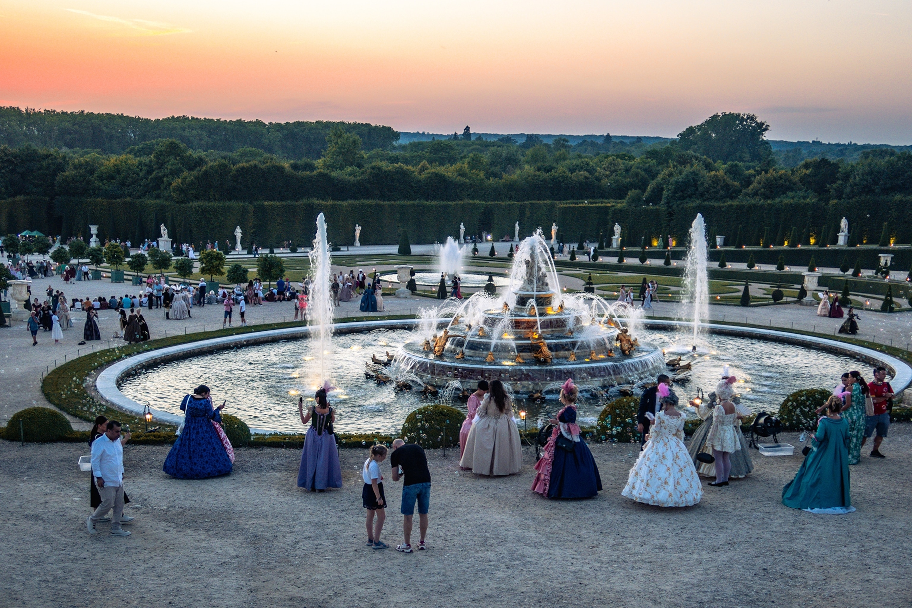 Guests dressed up in rococo-style dresses and frocks touring a royal garden and chatting around a fountain.