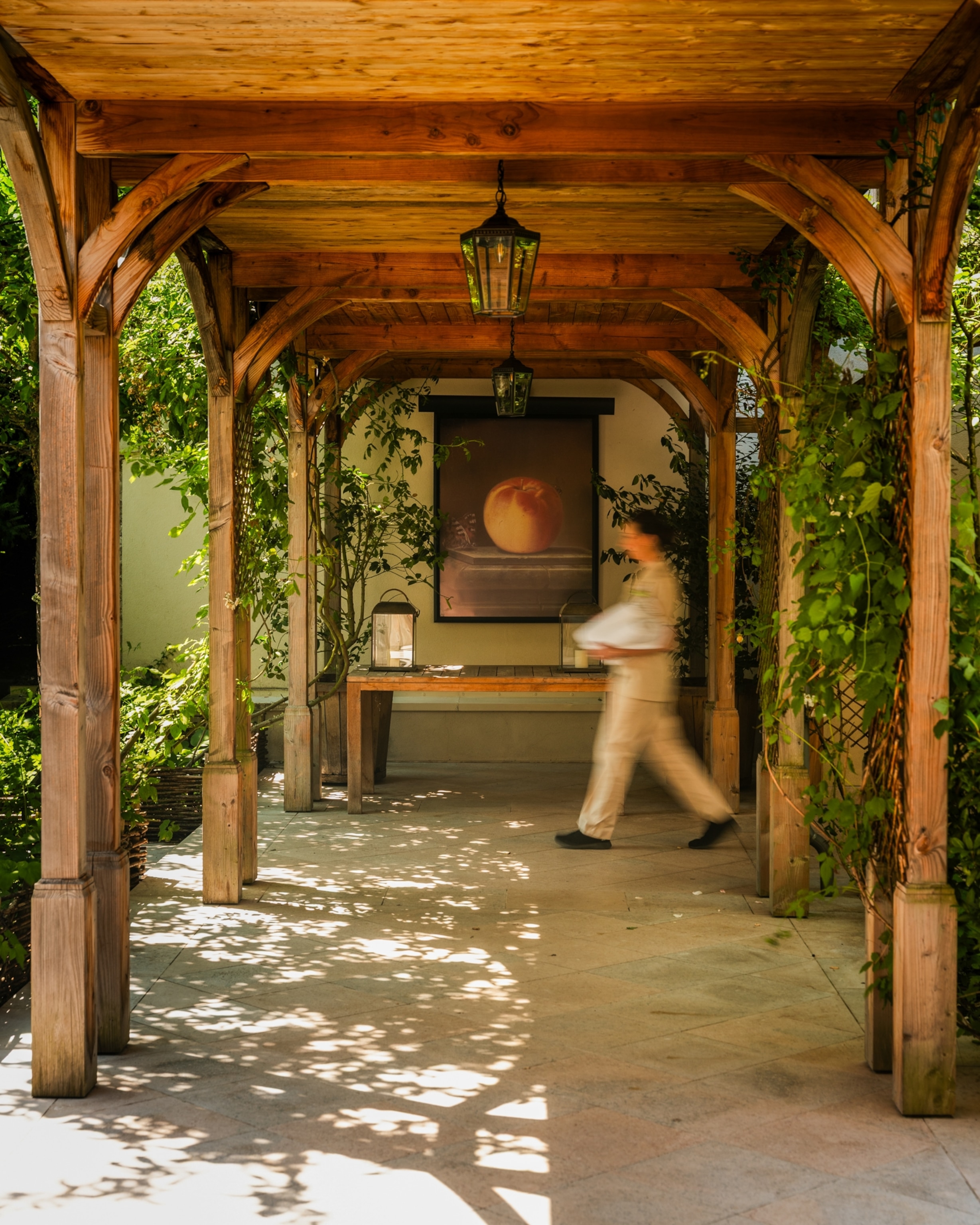 A wooden covered walkway lined with climbing plants leads to a seating area, while a person walks past a framed artwork at the far end.