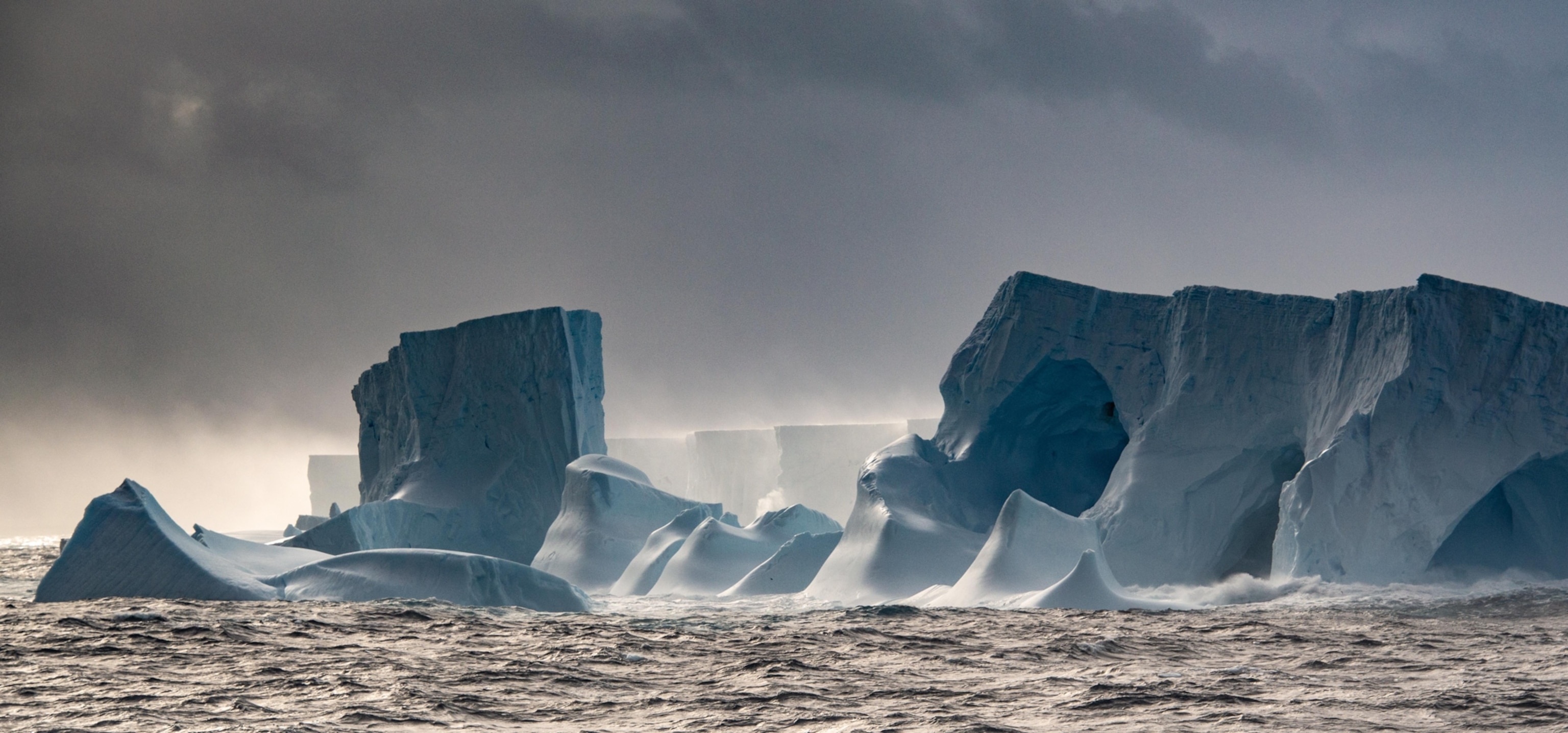 Massive icebergs float in a turbulent ocean under a brooding, cloudy sky