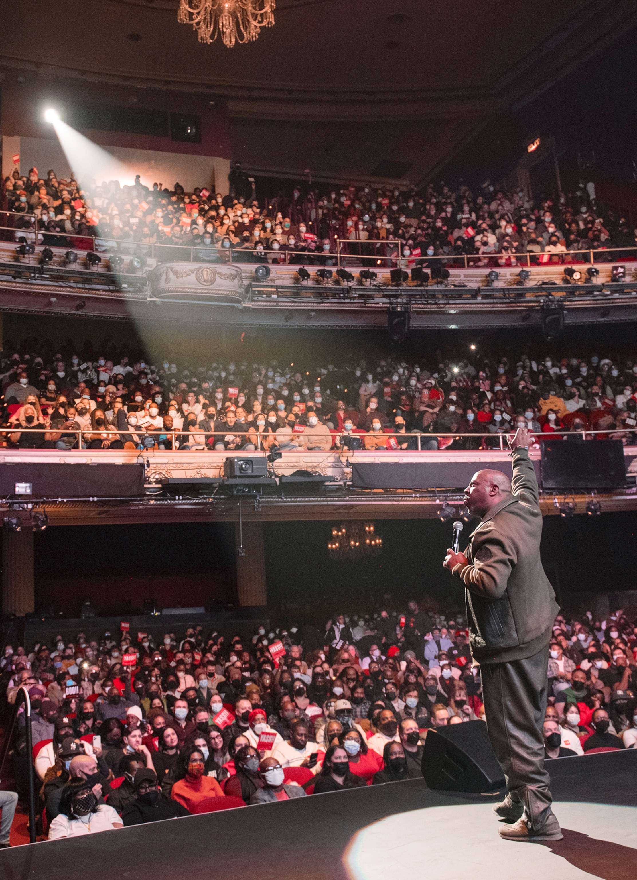 A man holding a microphone stands on a stage looking into a fully-filled crowd as a spotlight illuminates him.