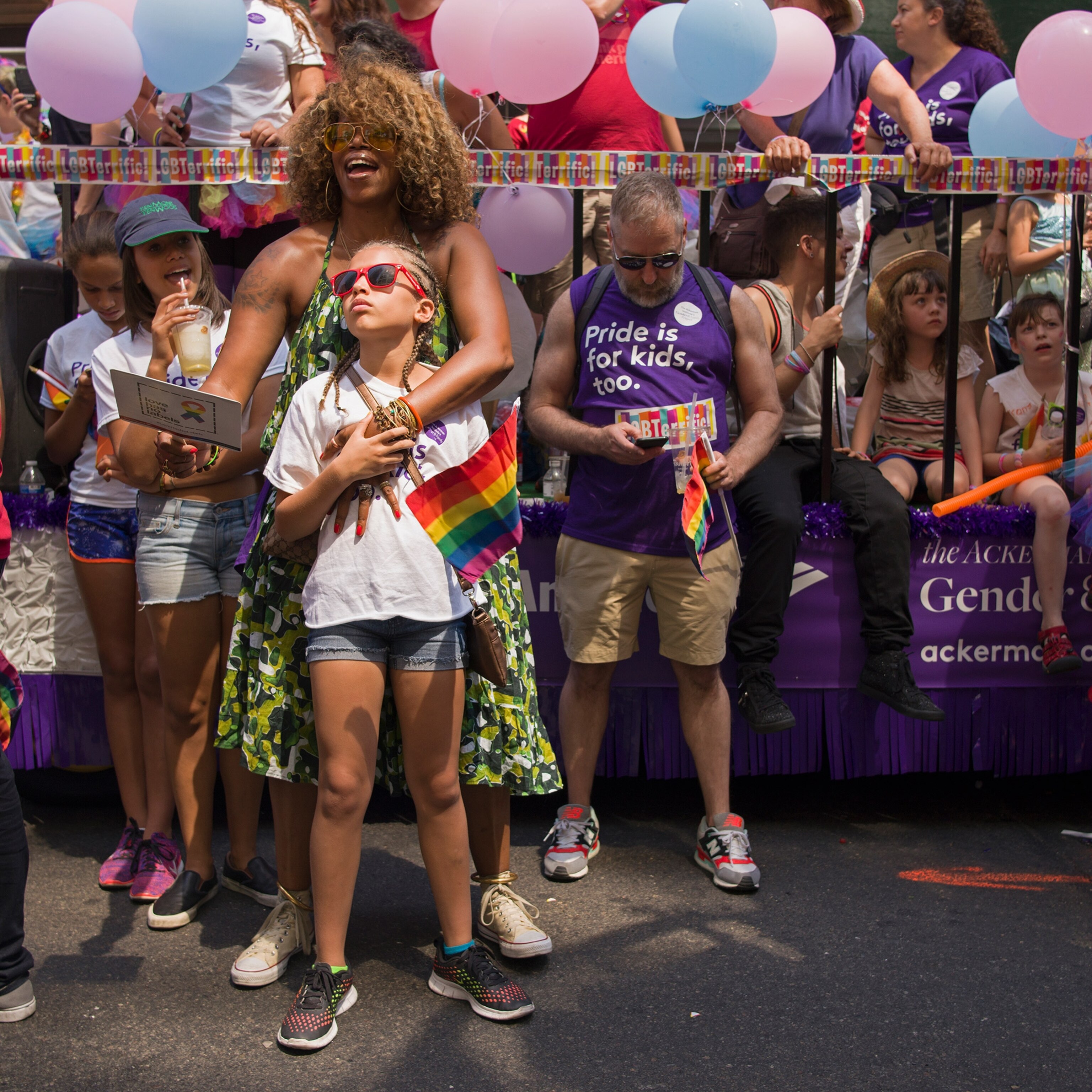 A child stands with their mother with braids and sunglasses during a parade.