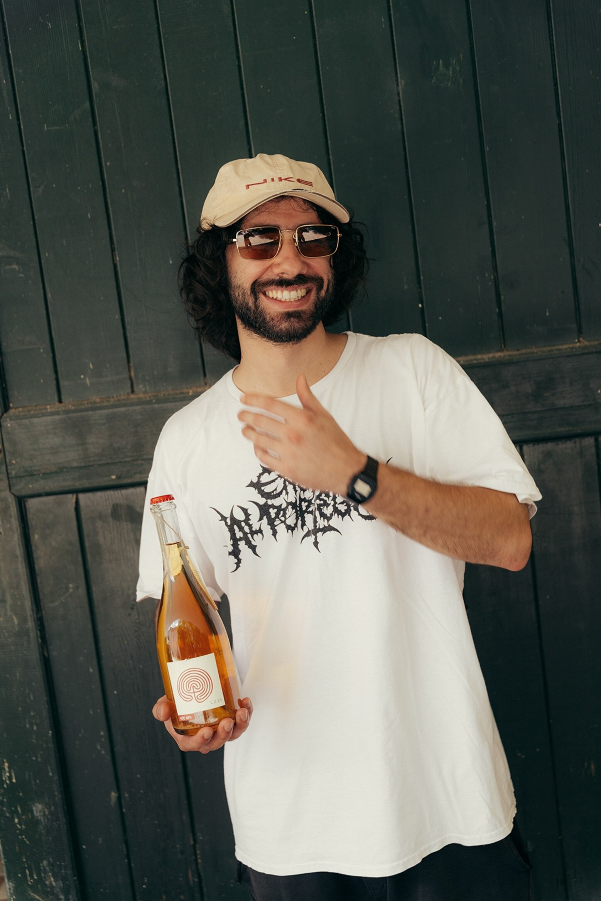 A young vintner holding a glass of prosecco, wearing a cap and t-shirt.