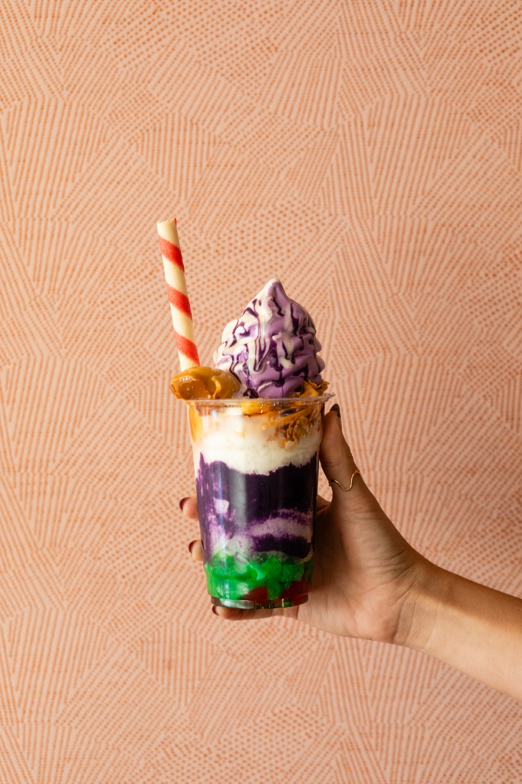 A hand holding a cup of haluhalo desert, which included layers of purple, green, yellow, and white.