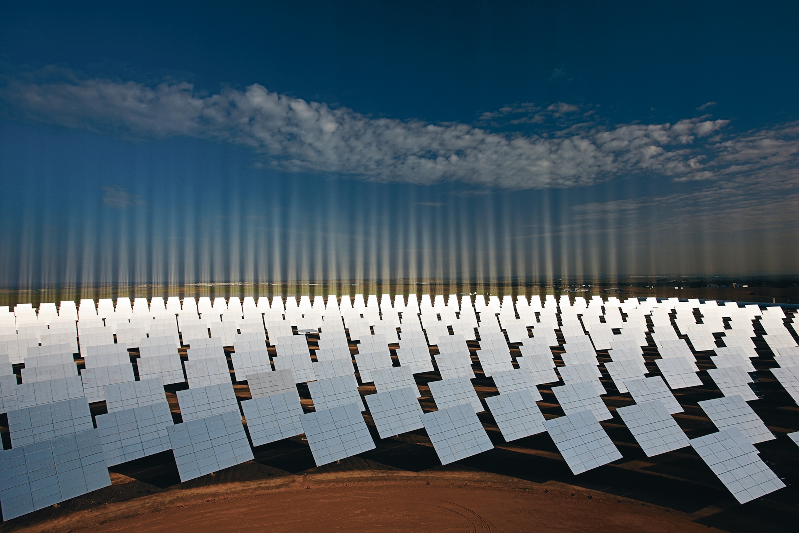 Rows of reflective solar panels stretch across a vast field under a clear blue sky with scattered clouds
