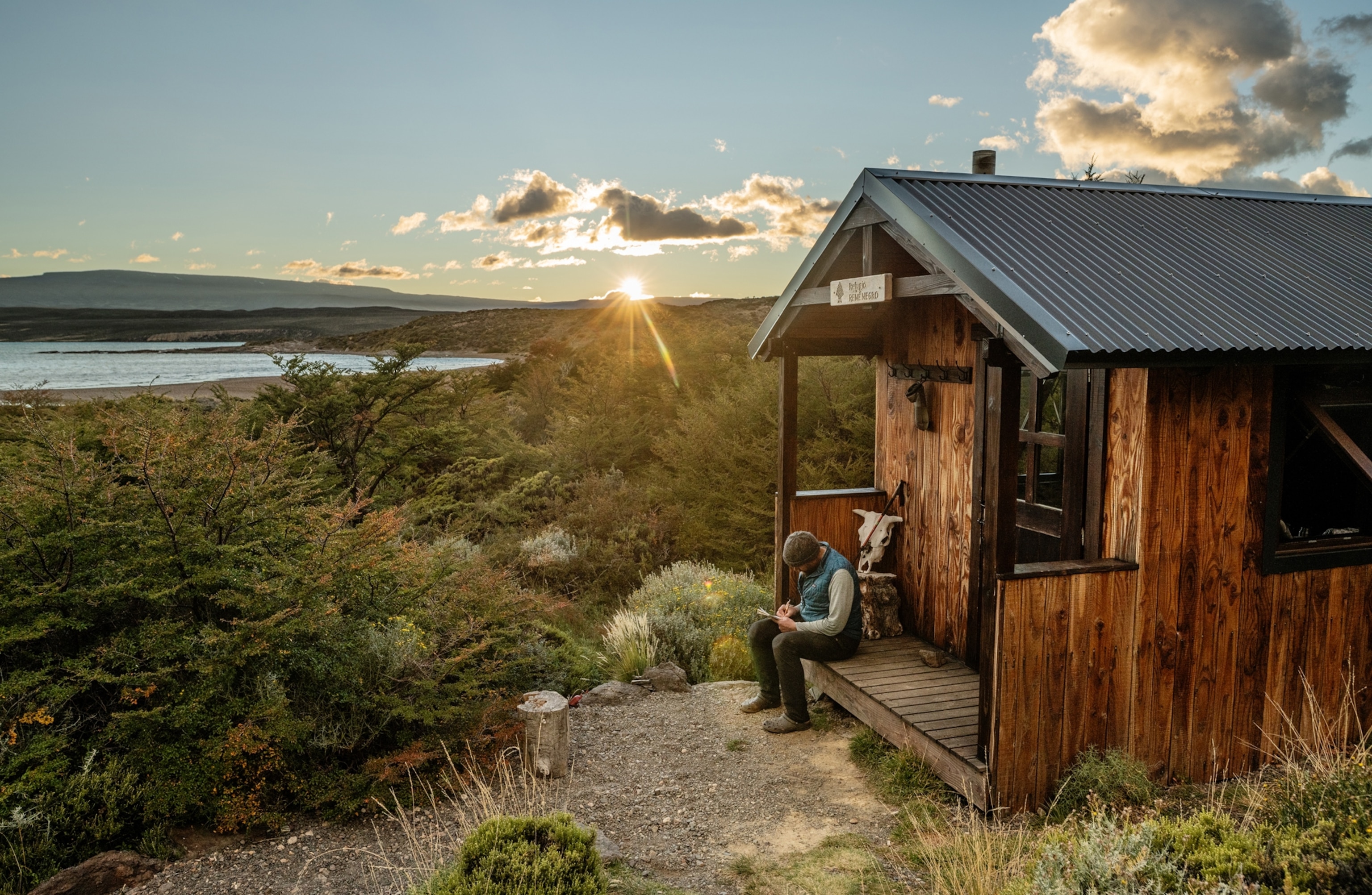 A man sits and writes in a journal on the front porch of a small cabin. There is a lake and greenery in the distance.