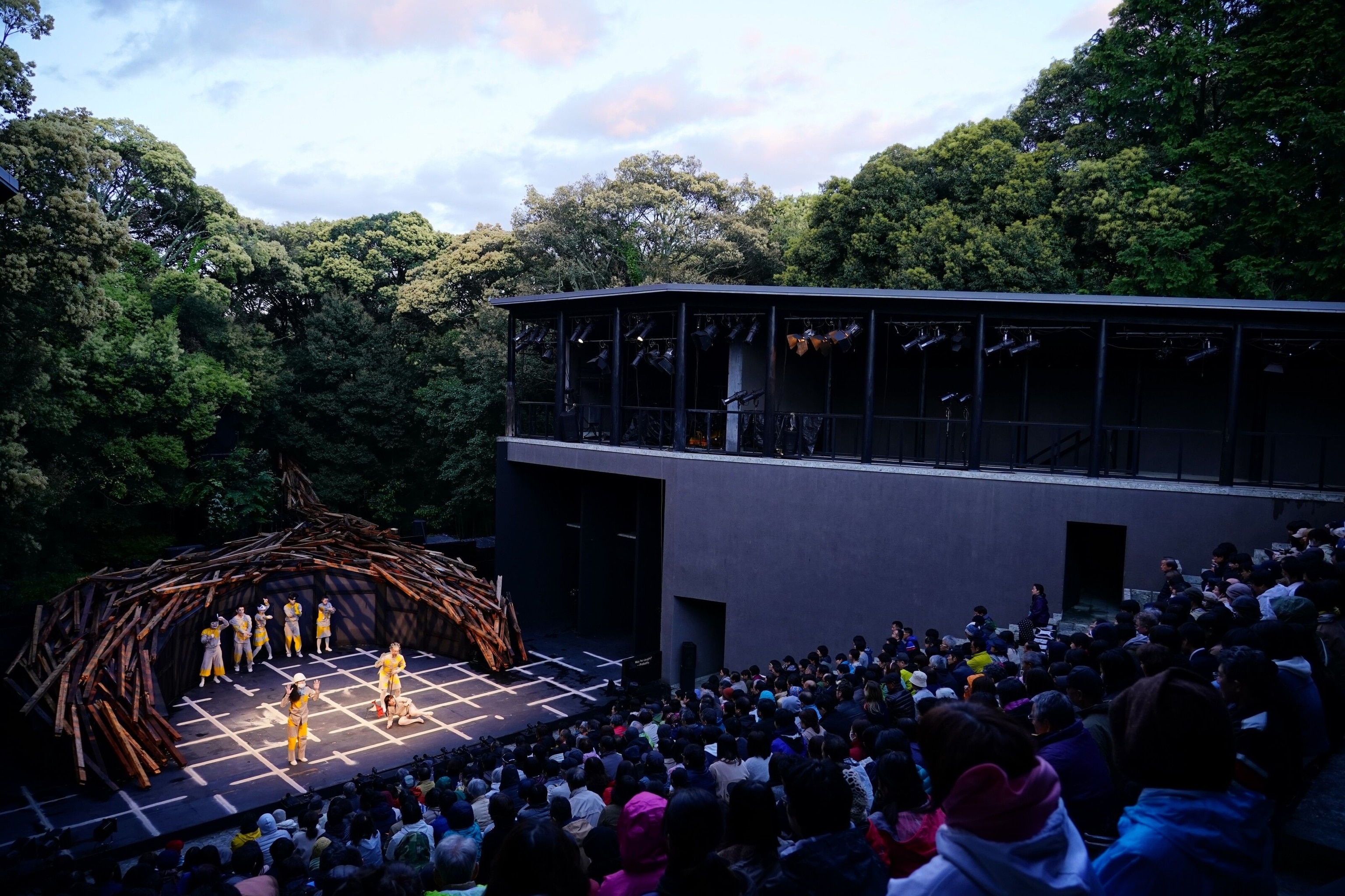 People performing in an outdoor theatre. The actors wear yellow and grey outfits and the stage looks like a willow arch without any greenery. There is a large crowd, the sky is blue but spotted with clouds.