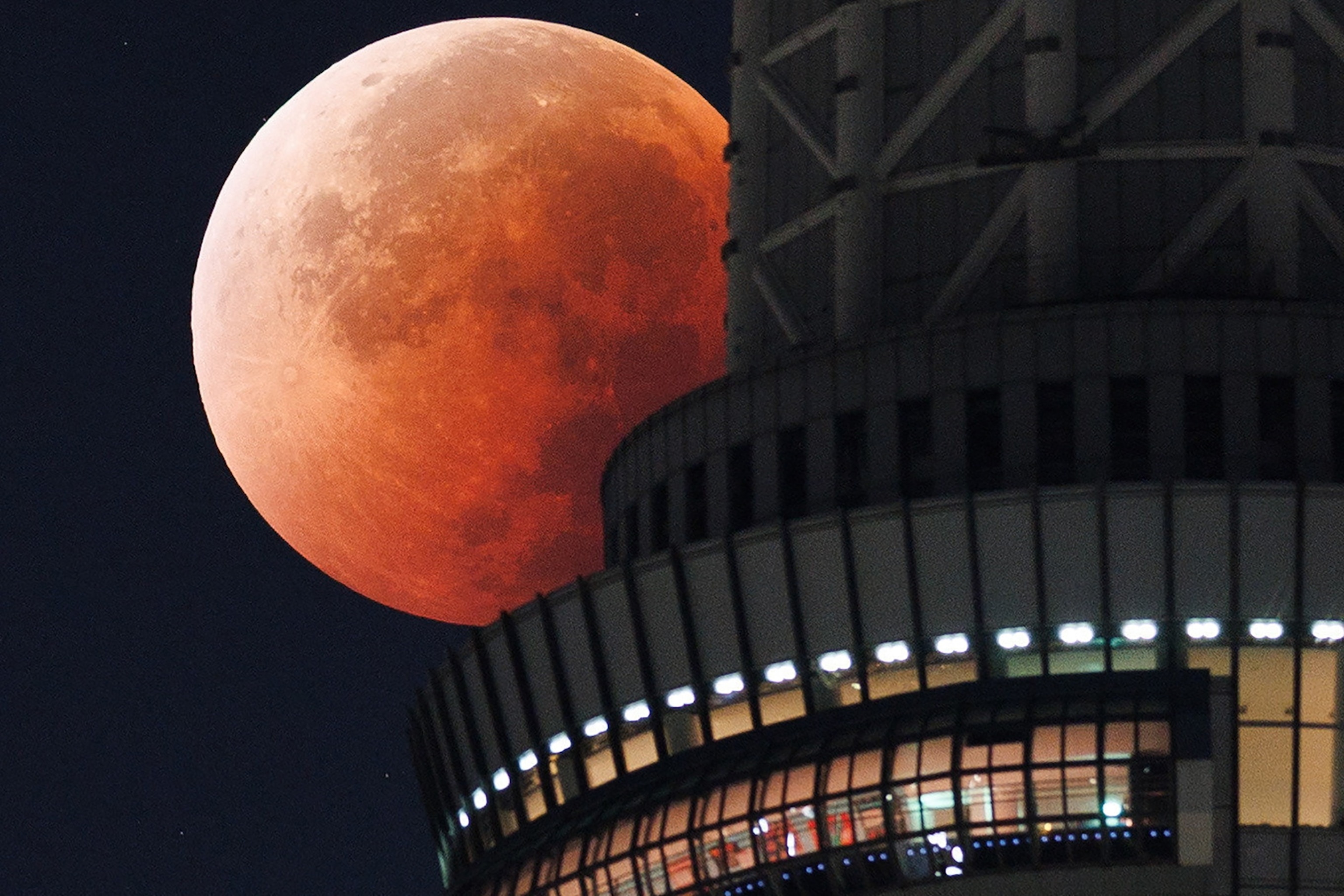 A large, reddish moon during a lunar eclipse is partially obscured by a modern, cylindrical skyscraper at night
