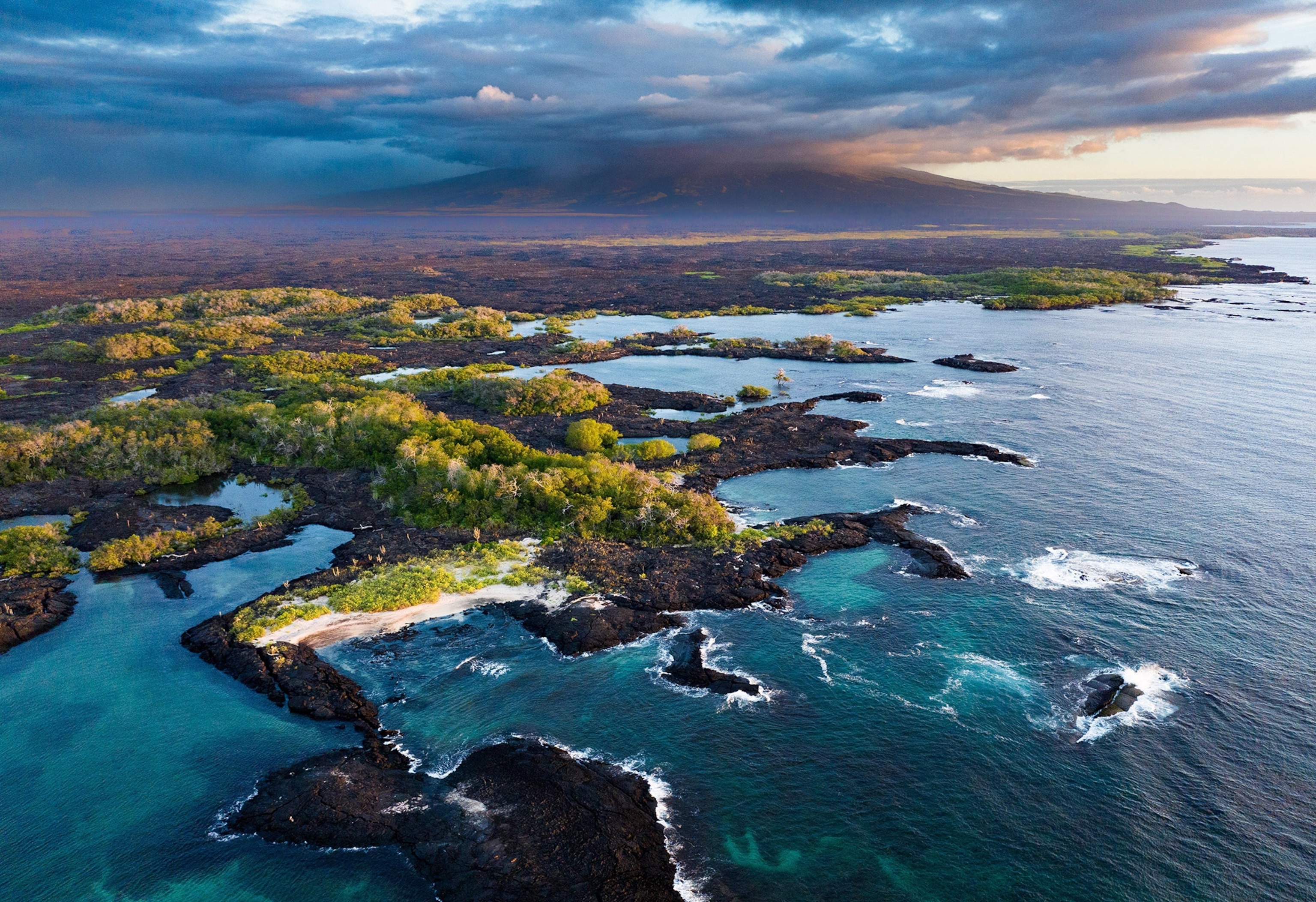A narrow land mass, topped with greenery and lined with black volcanic shore, juts out into a dark blue ocean. Sun peers through dark clouds from the right.