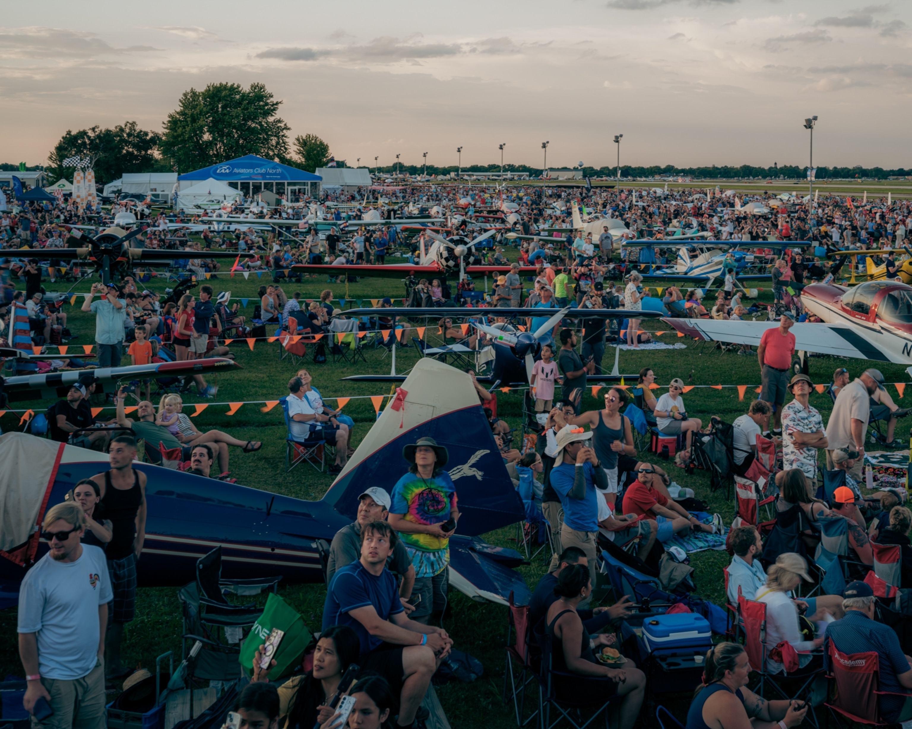 A large, lively crowd gathers on a grassy field at an airshow, surrounded by small aircraft. People are seated on chairs, talking, and enjoying the event.