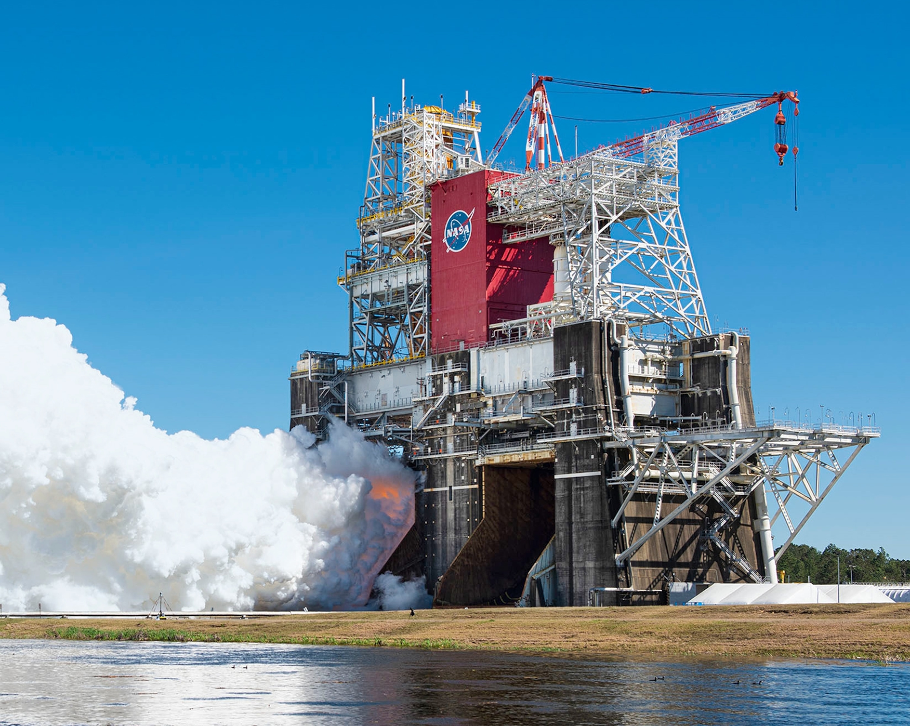 A large metal structure used to test rockets sits on land behind a body of water and white smoke is coming out of its base.