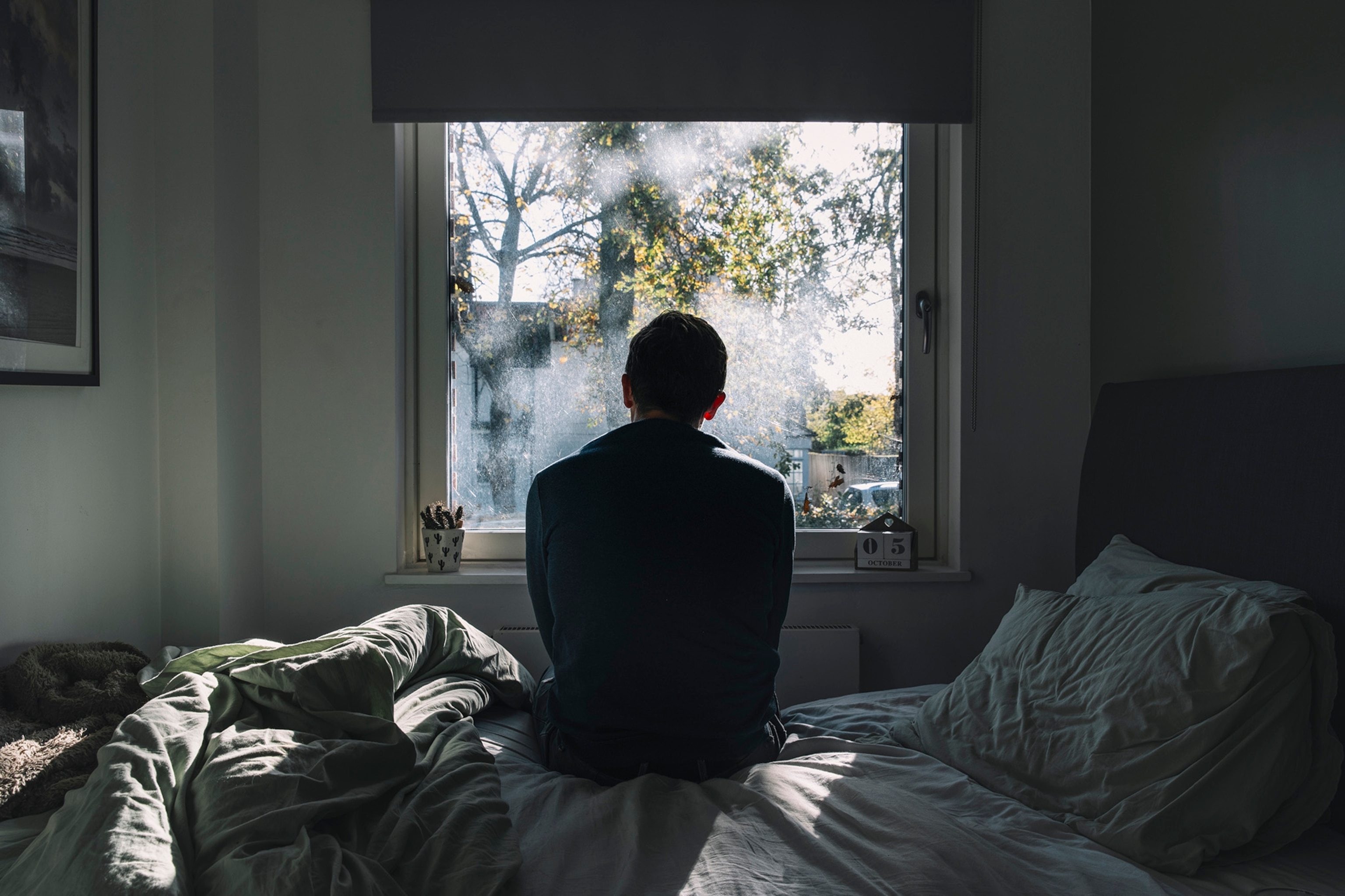 A man sits on a messy bed facing a window where sunlight streams through trees.