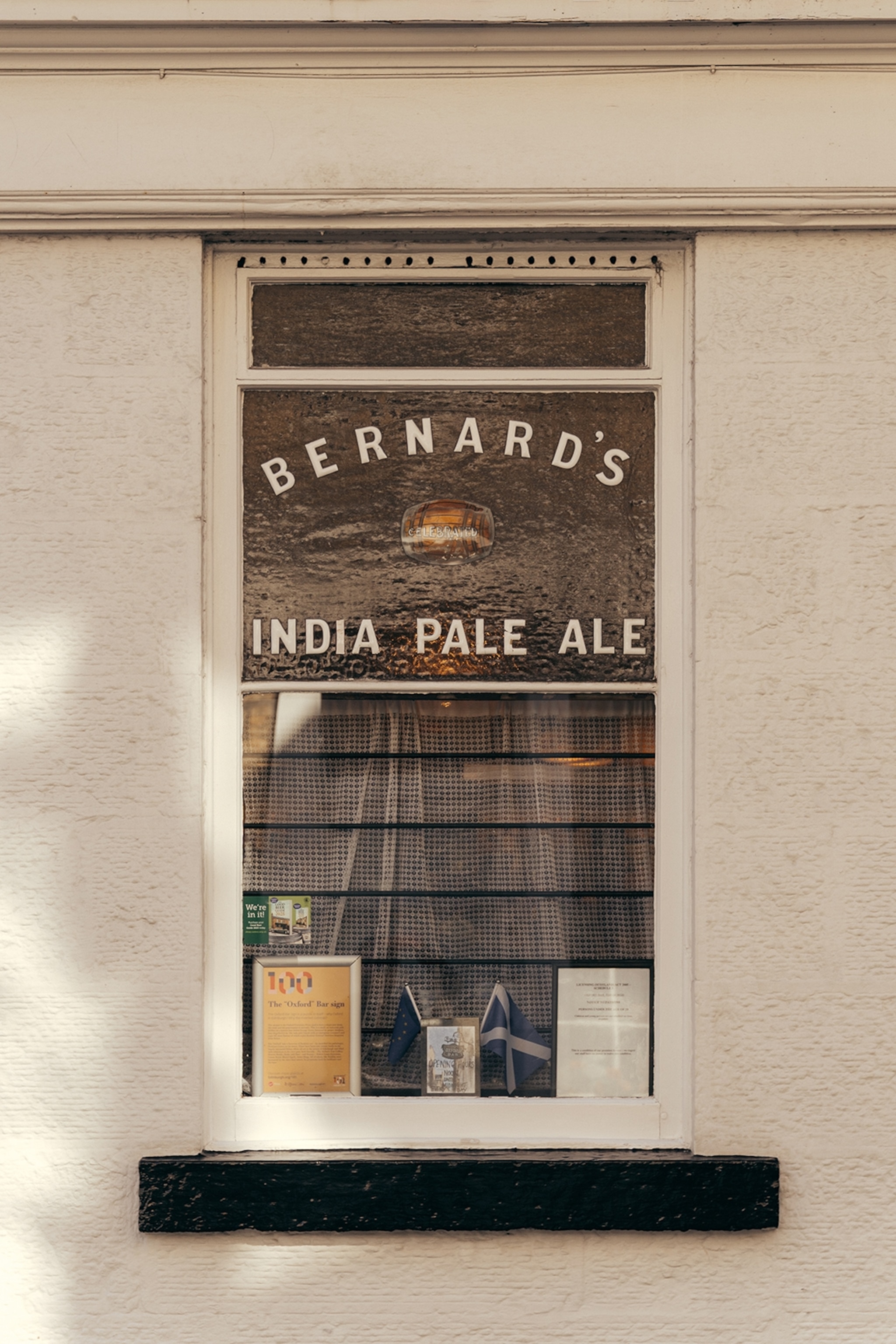 The exterior window of a simple pub with cafe curtains and 'Bernard's Indian Pale Ale' written on the glass.