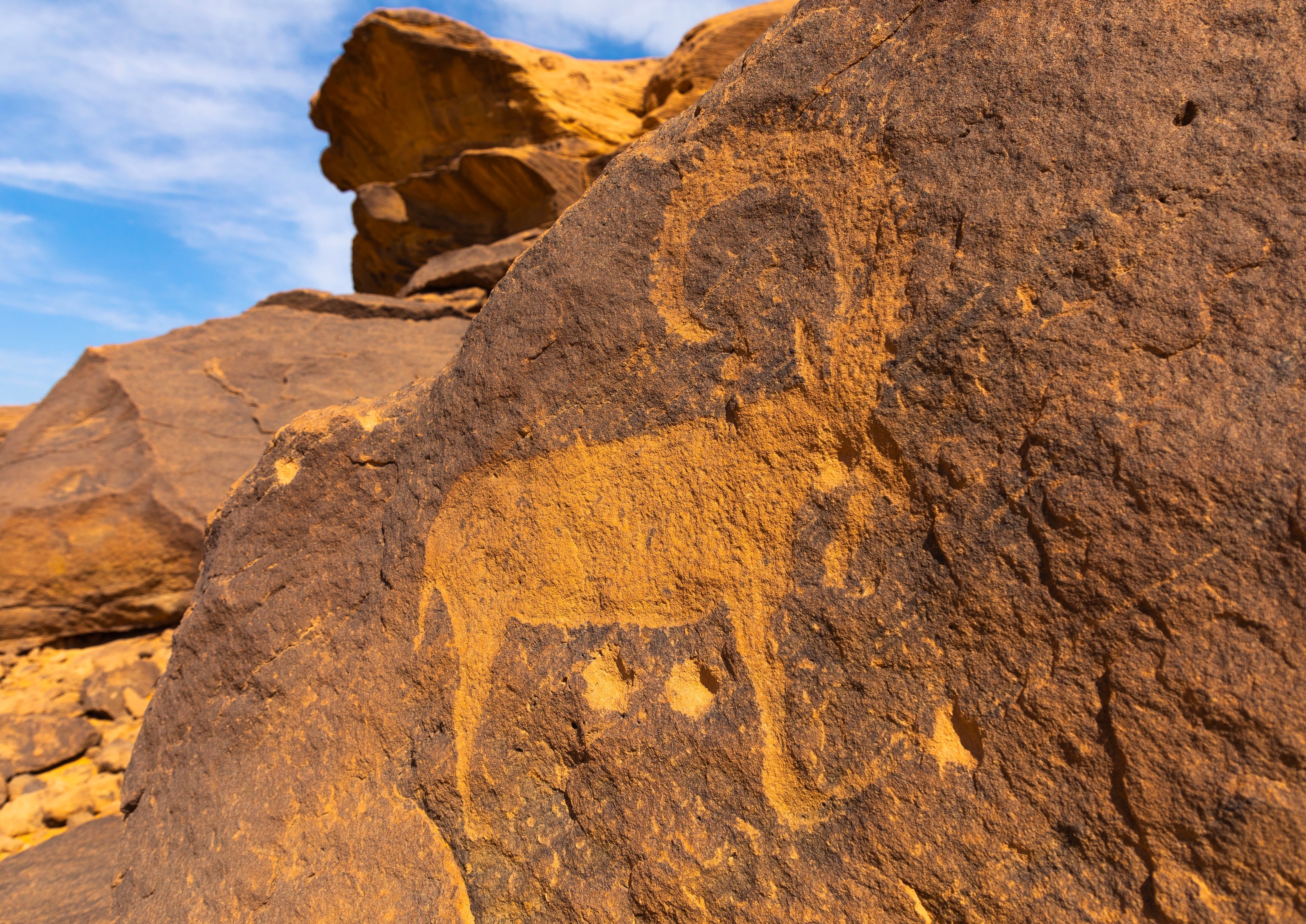 An ancient rock carving of an ibex is etched into a large boulder in a desert landscape of weathered sandstone formations.