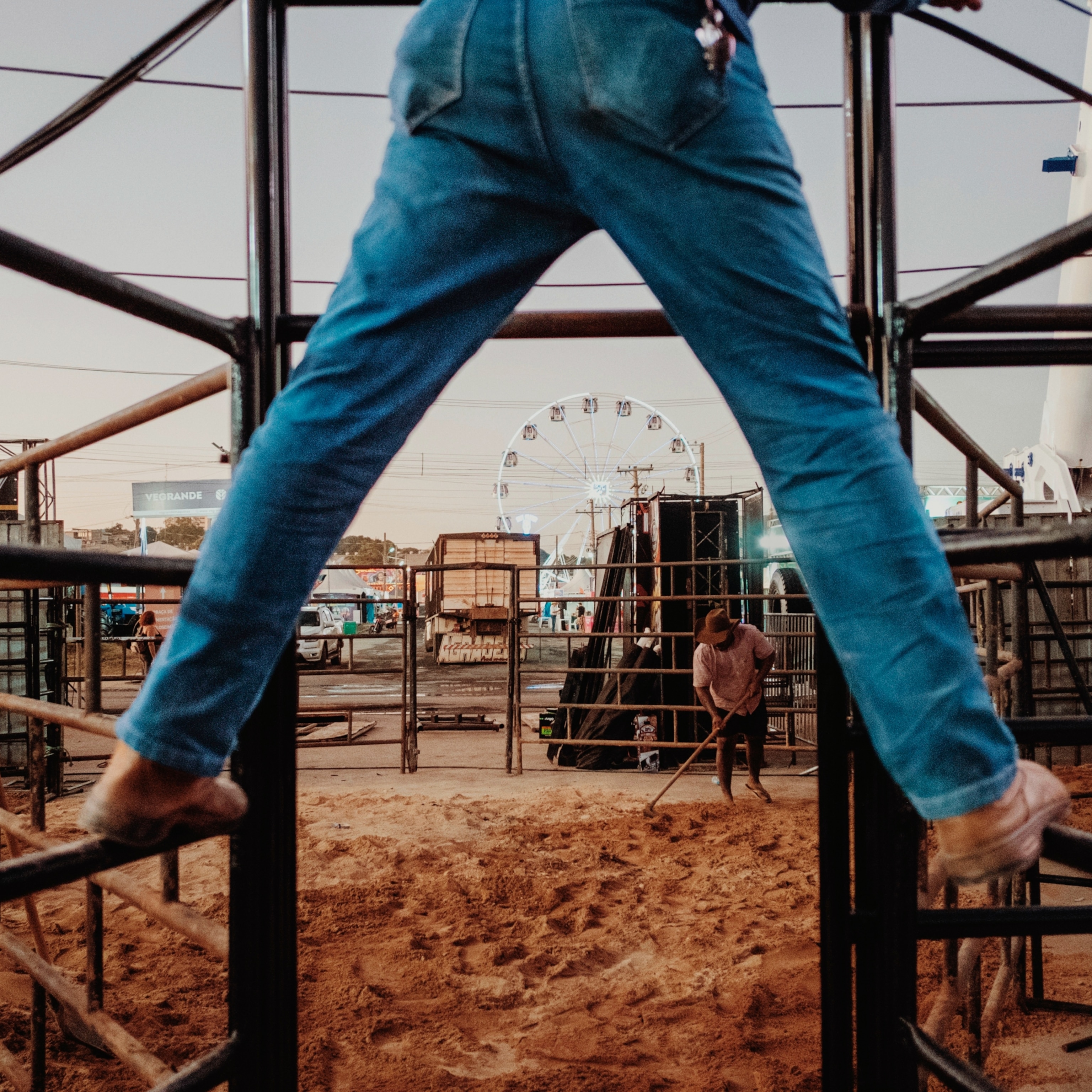 A pair of legs in blue jeans standing on a bull gate in front of a ferris wheel.