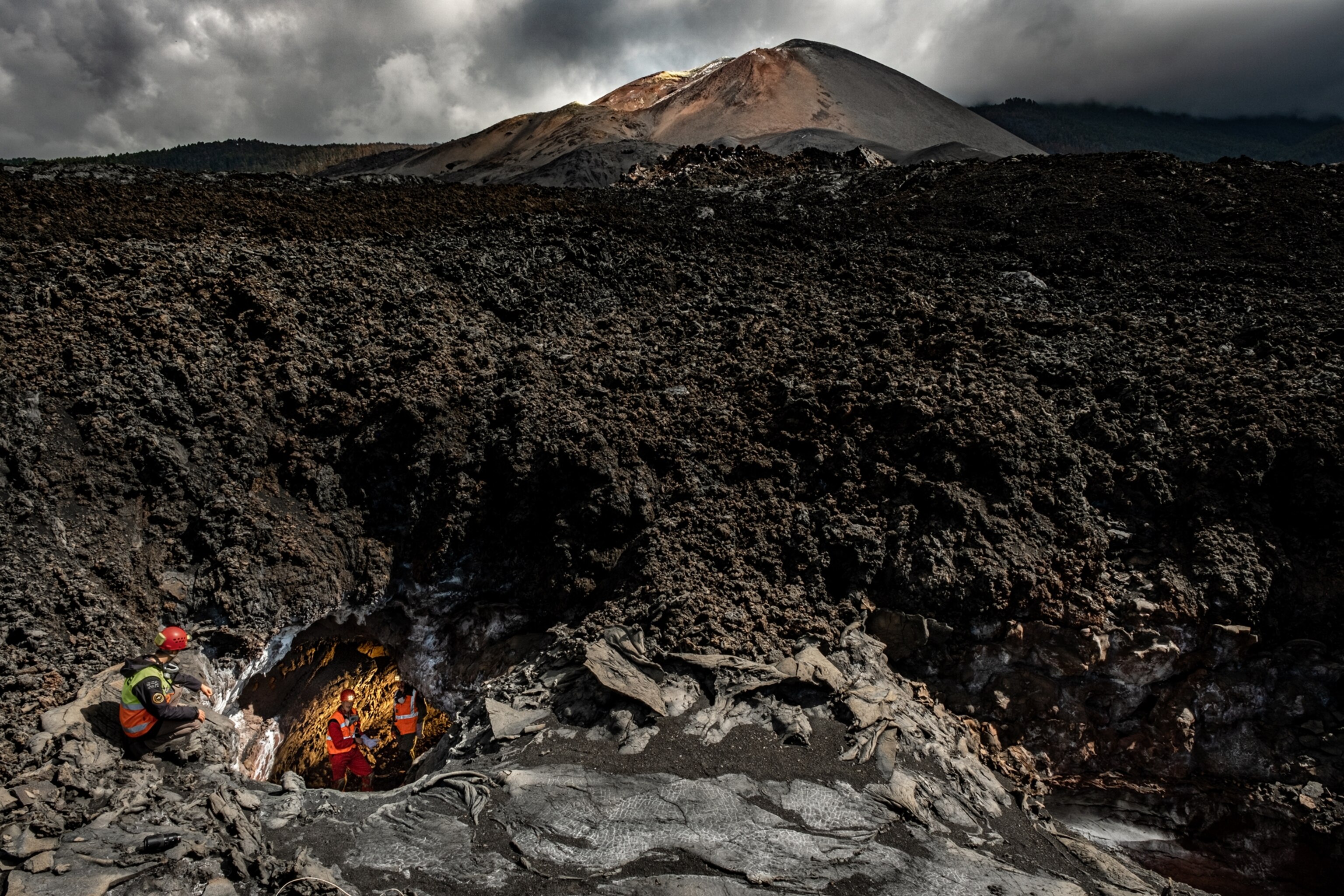 Tajogaite volcano standing before an enormous field of black ash and lava rock; in the foreground, three researchers in orange safety vests are examining the entrance to a lava tube that appears to run beneath the field of lava rocks.