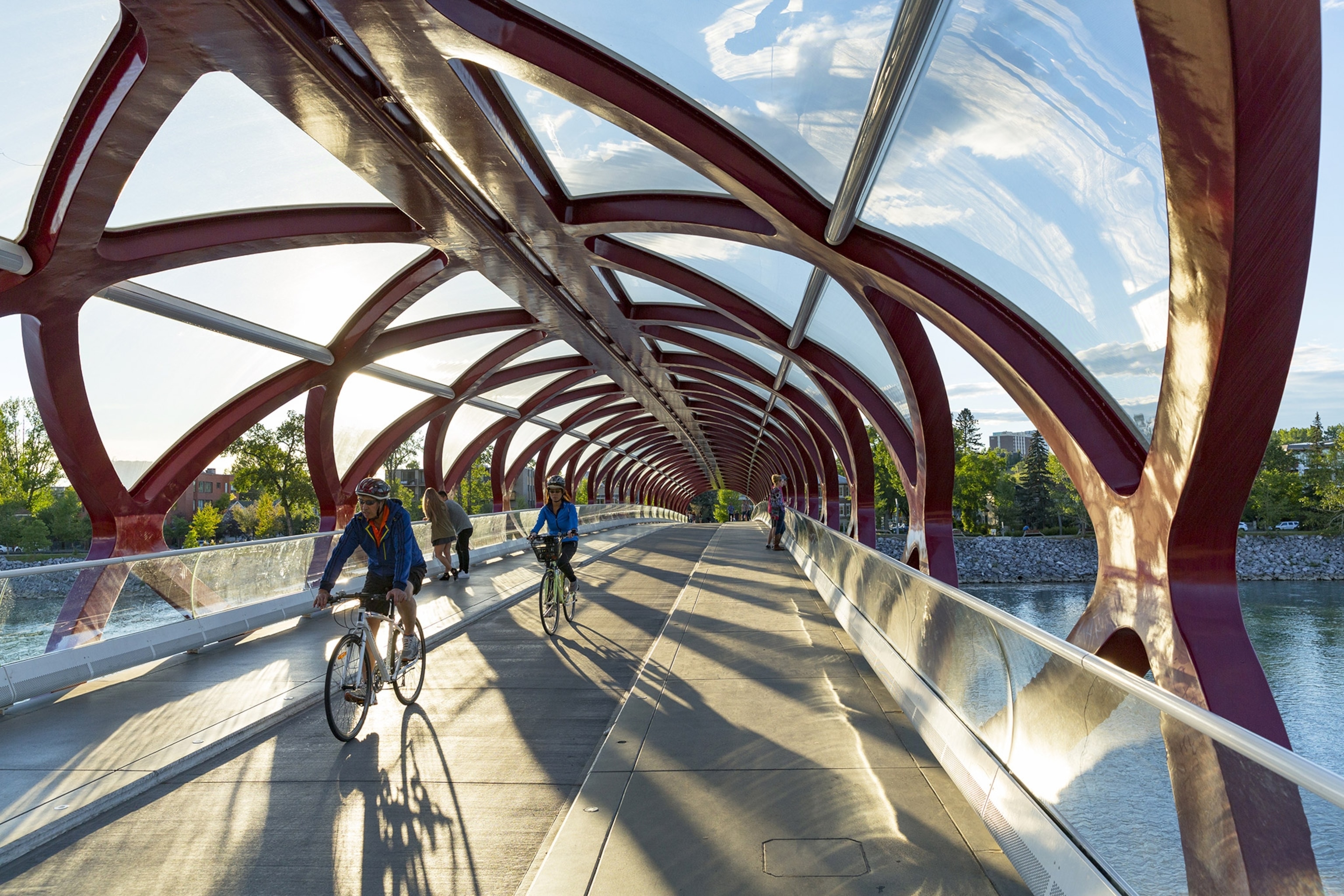 Cyclists ride toward foreground along a straight tube-like bridge covered with red geometric covering