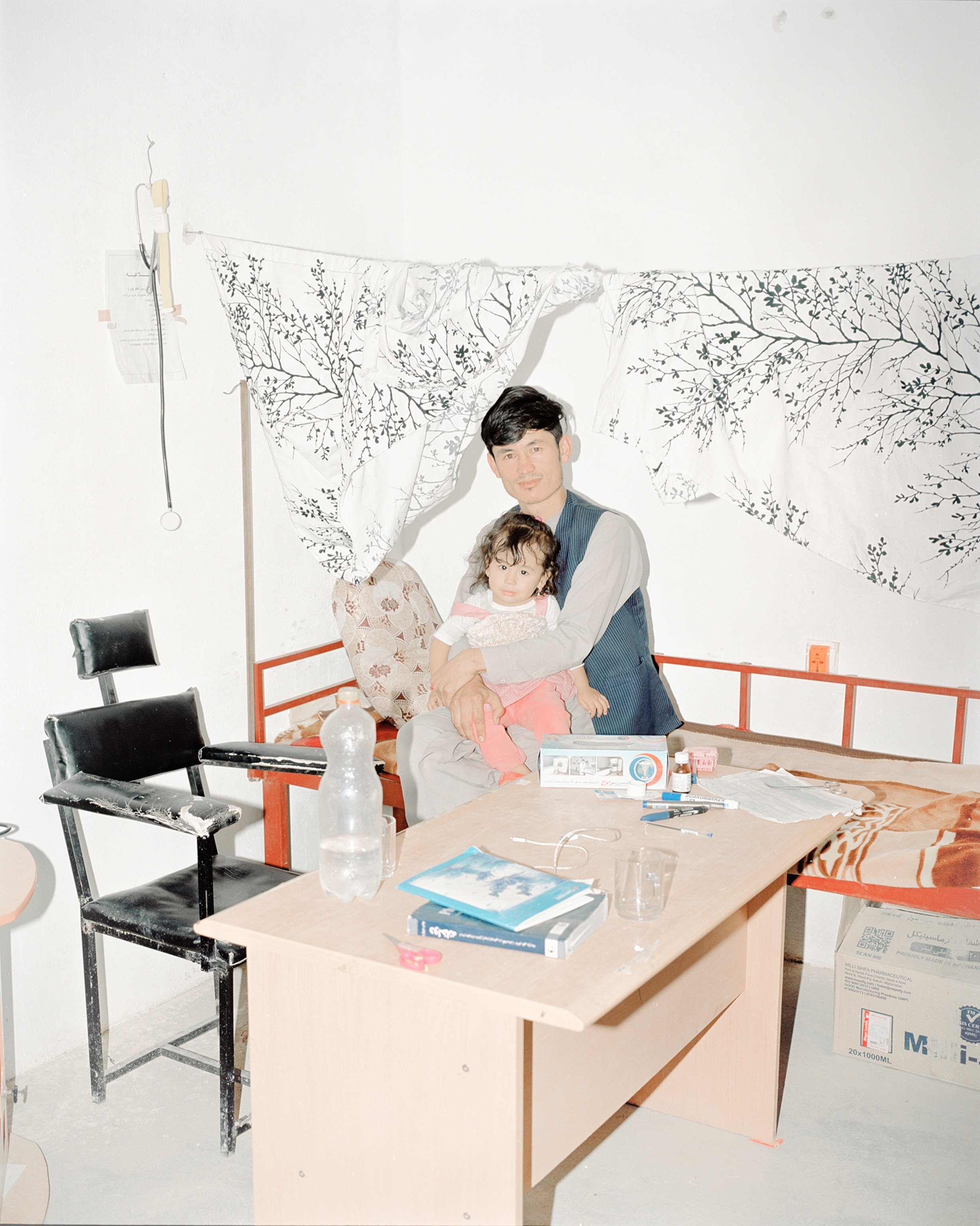 A man sits on a bed with his young daughter on his lap with a desk and chair in front of the pair.