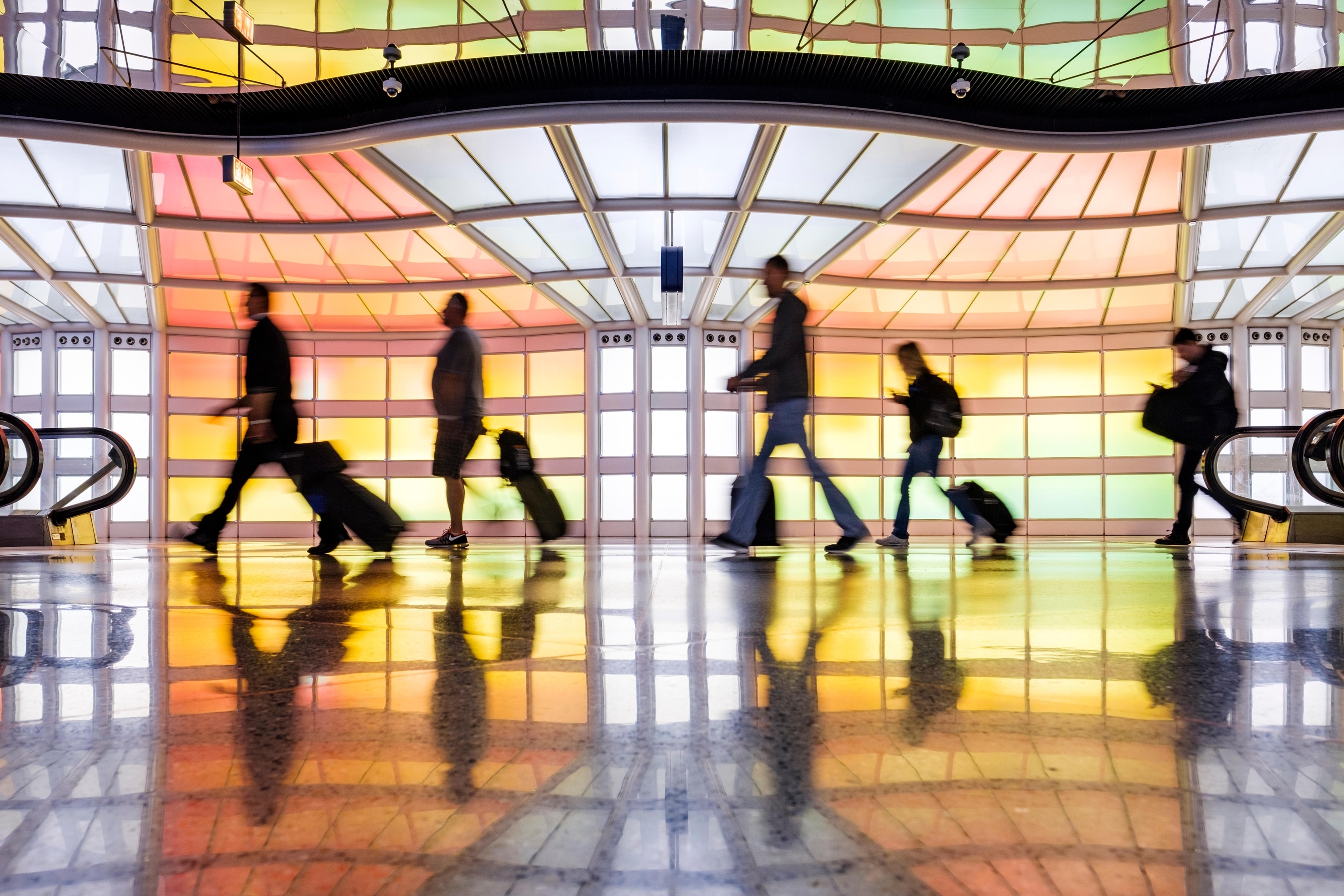 Side view of silhouetted pedestrians pulling suitcases in front of an airport wall featuring neon-lit panels of alternating colors.