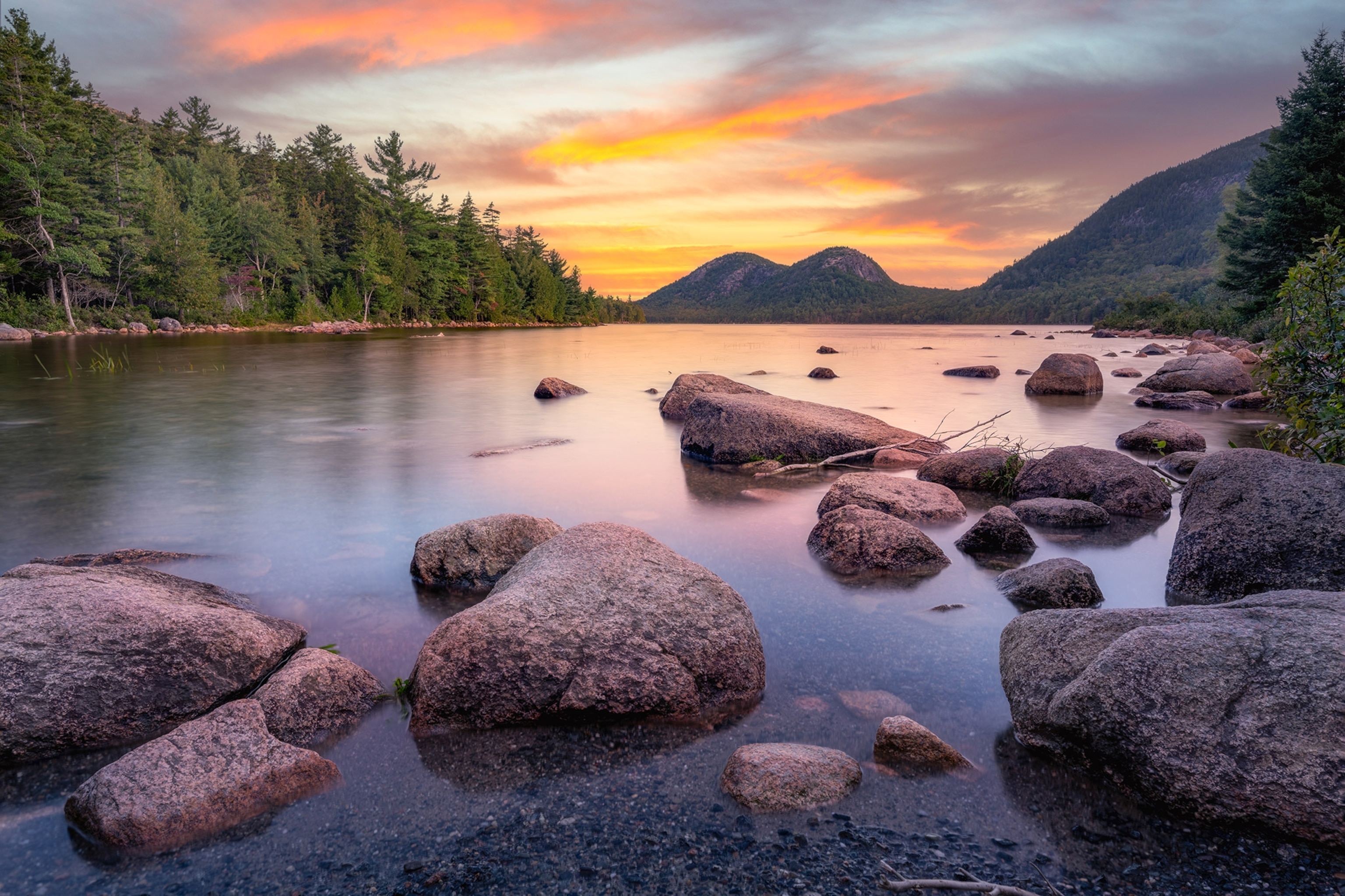 An orange and pink sunset is reflected in a still pond surrounded by trees and mountains, round rocks in the foreground.