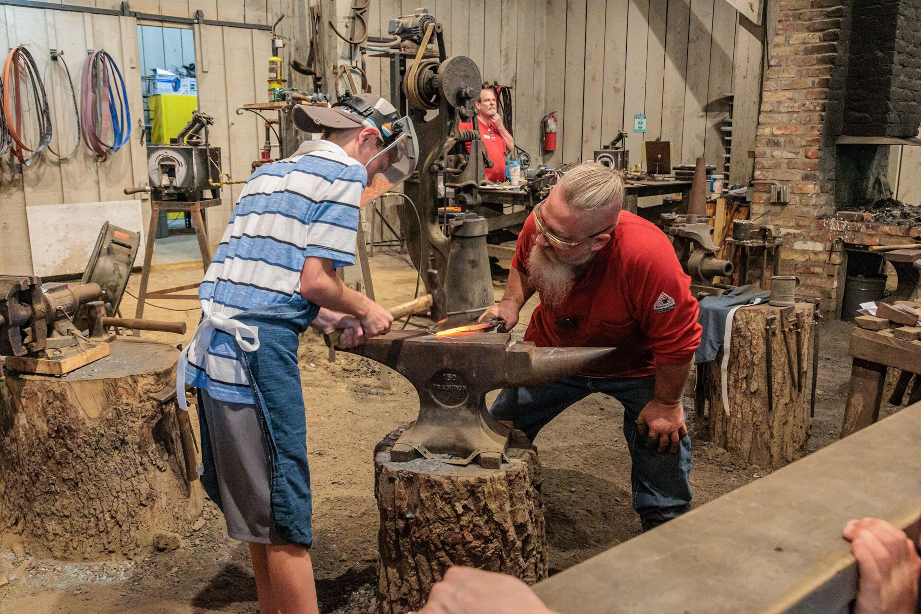 A boy wearing protective face mask wields a hammer over a glowing piece of steel on an anvil, a man on the other side bends down to oversee.
