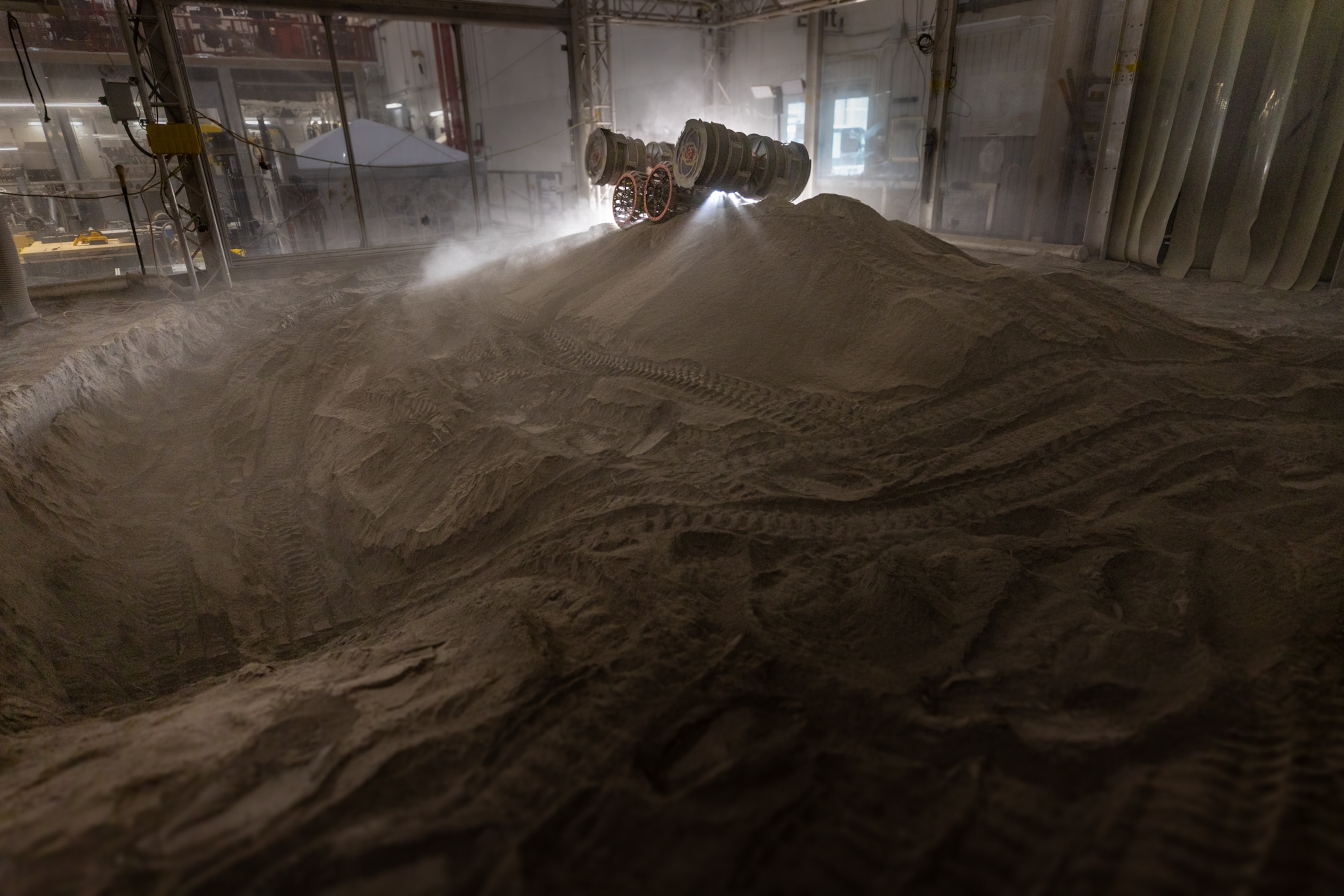 A lunar rover navigating a large indoor dune terrain, kicking up clouds of dust. The dimly lit, industrial setting creates a moody, adventurous atmosphere.