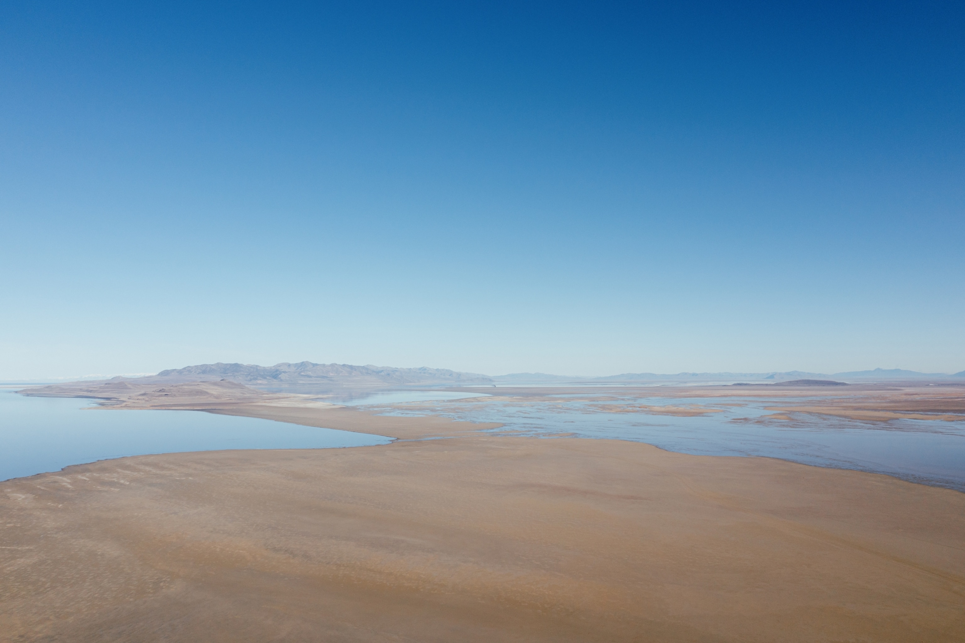 Aerial view of a vast barren landscape meeting the edges of a receding blue lake. Distant mountains are visible on the horizon.