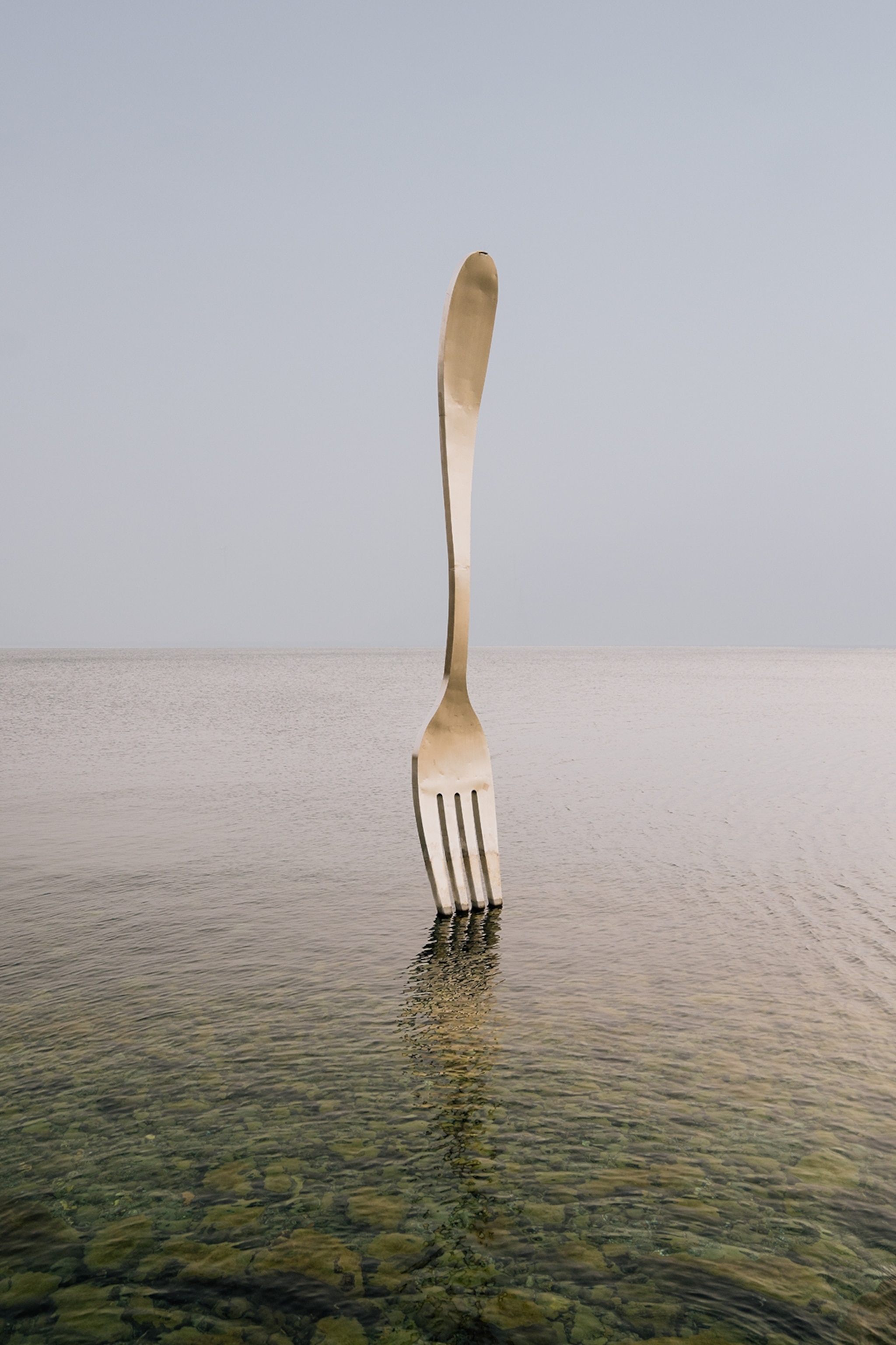 An oversized fork sculpture standing upright with its teeth down in a lake.