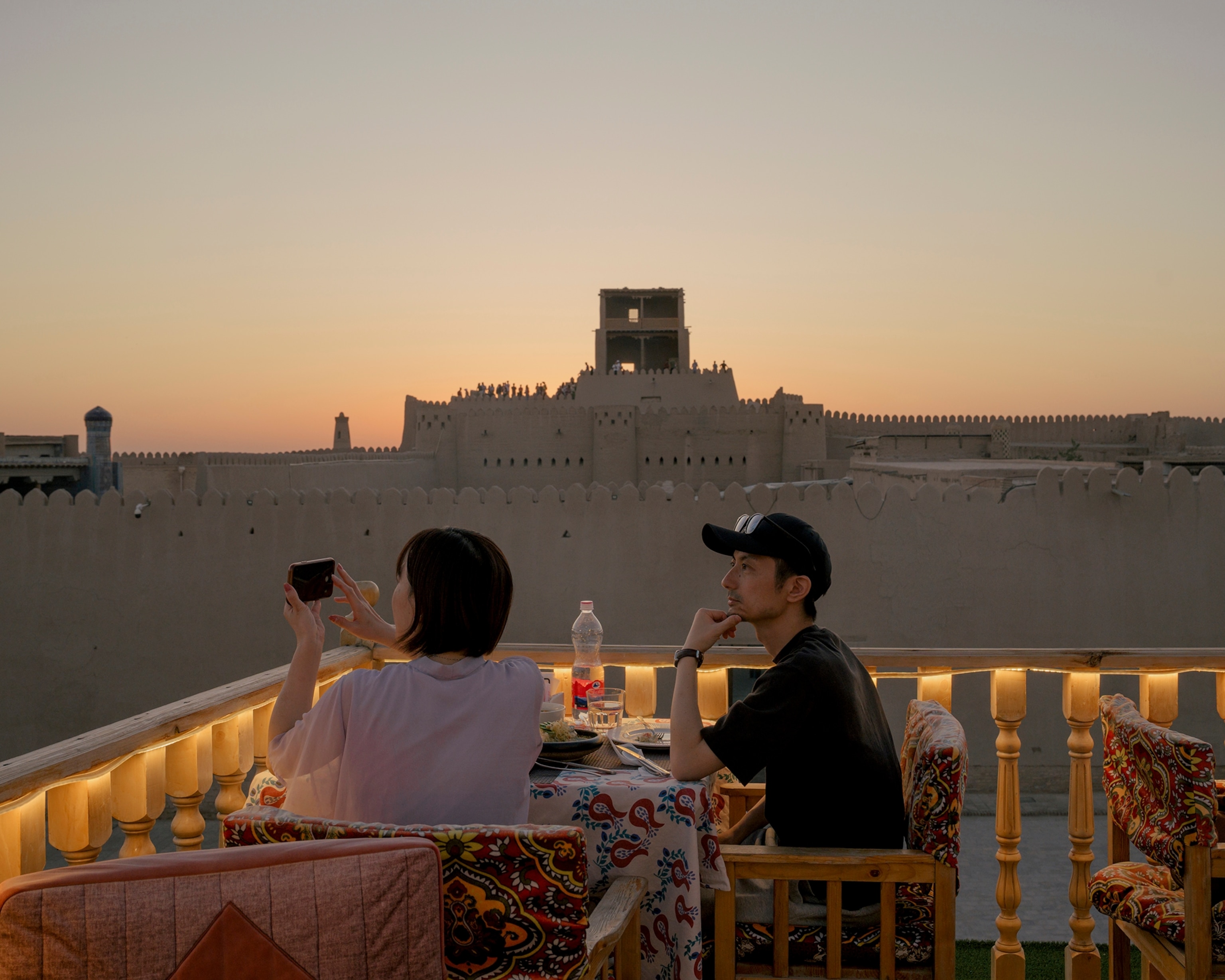 Tourists take photos during sunset on the rooftop terrace of the Terassa Cafe and Restaurant in Ichan-Khala, Khiva, Uzbekistan on August 15, 2025. The watchtower of Kuhna Ark can be seen in the background.