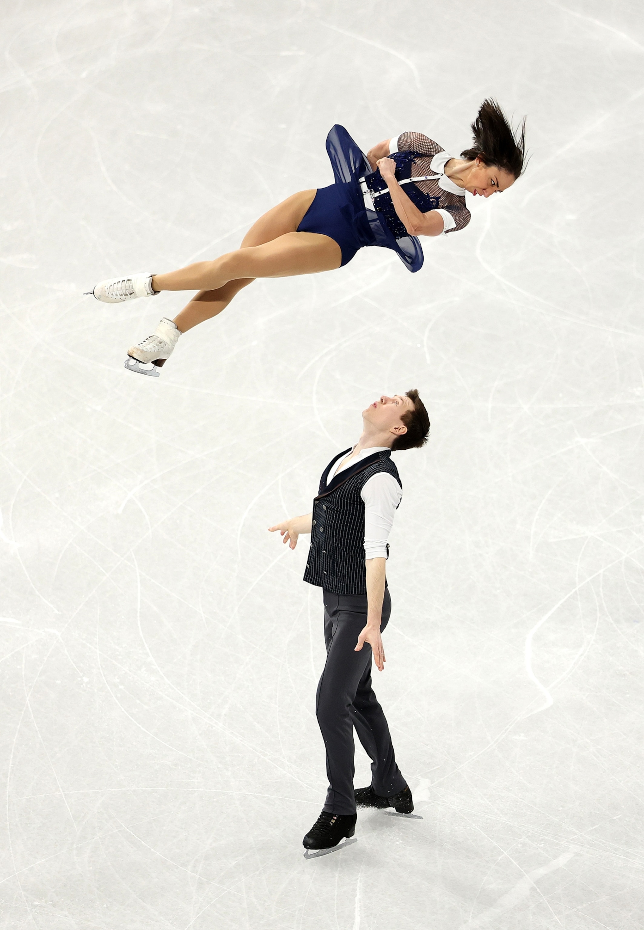 A woman spins in the air above a man who stands on an ice rink.