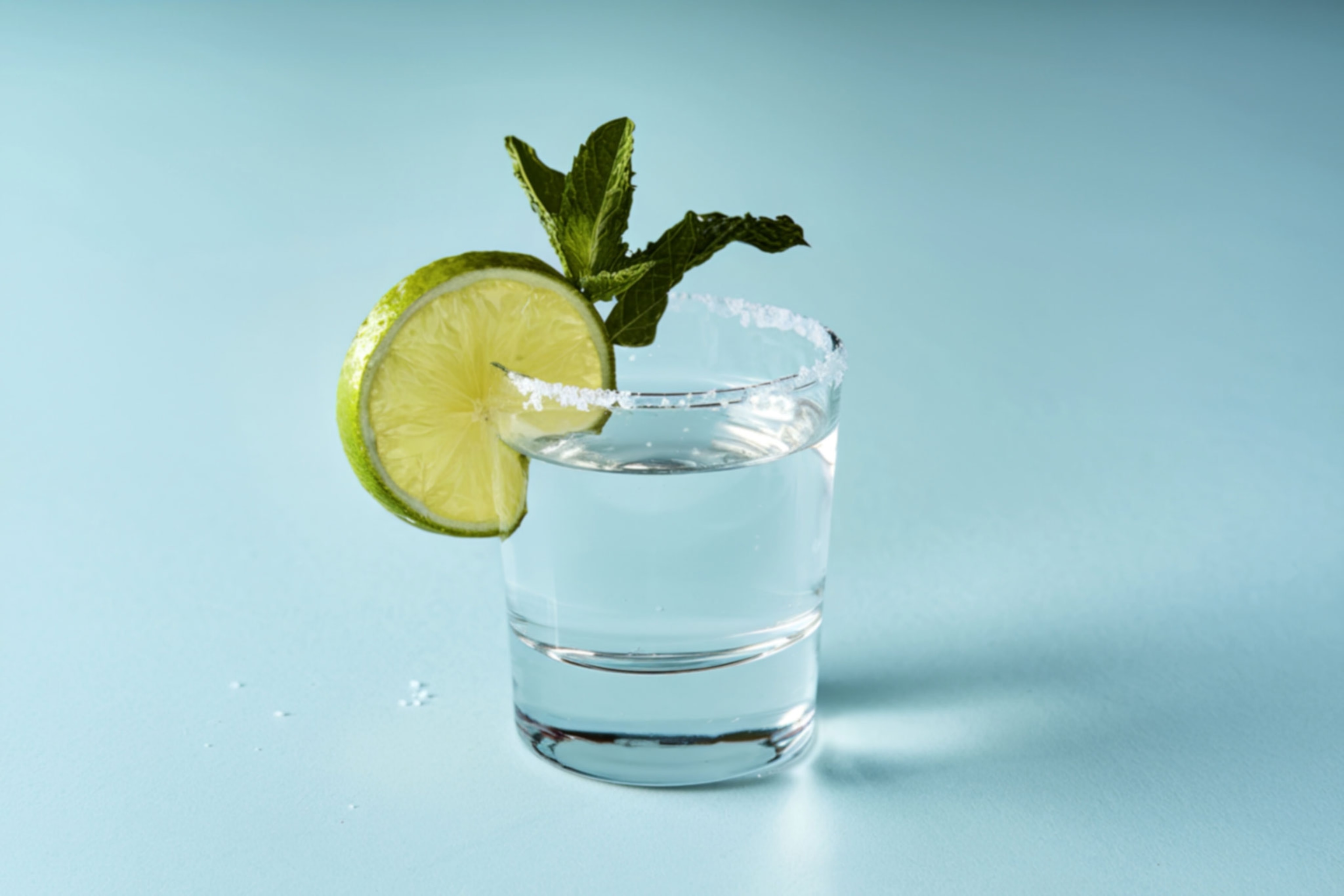a tequila shot with a lime wedge and salted rim and a sprig of mint against a light blue background