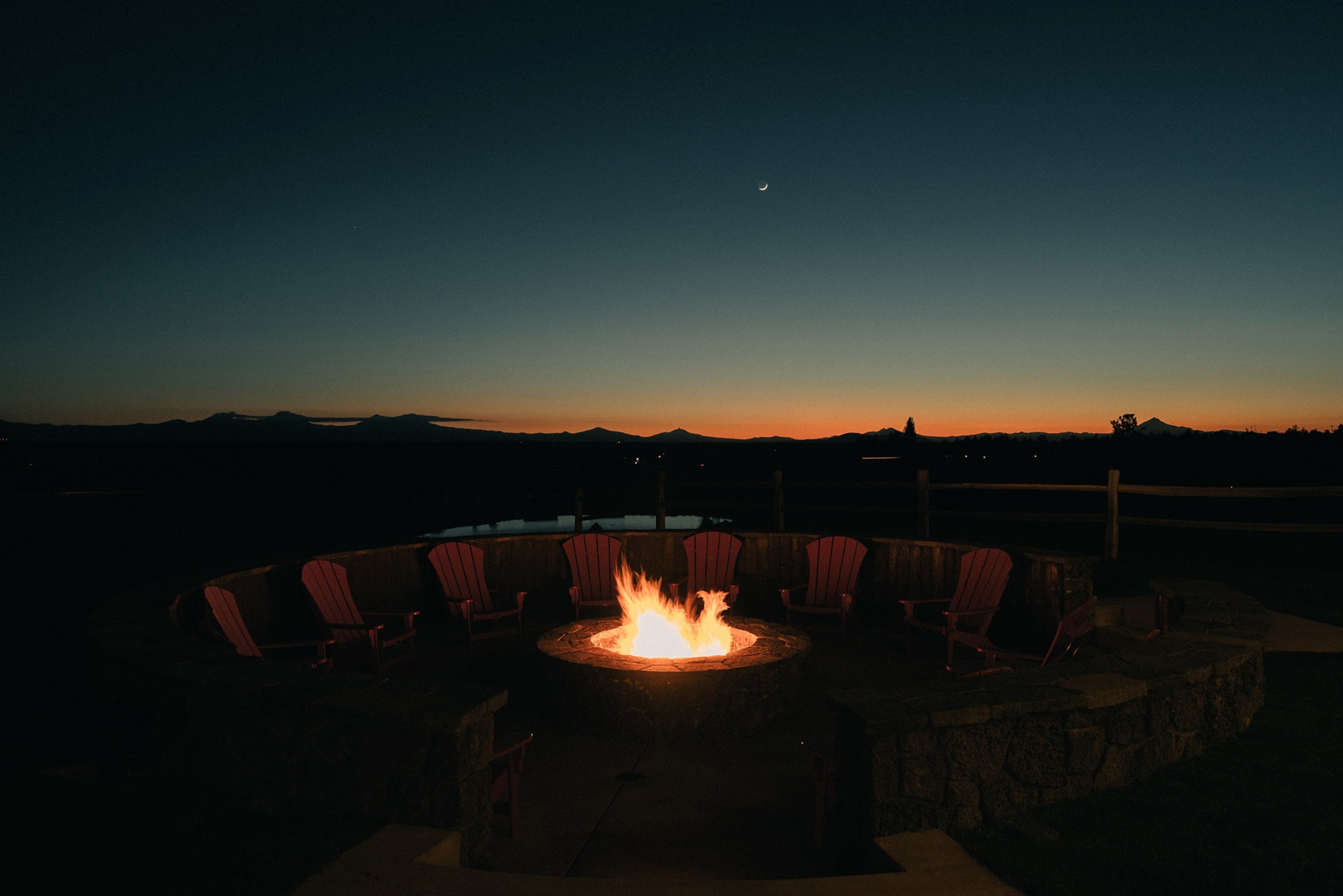A fire in the center of the frame surrounded by dimly-lit chairs arranged in a circle around it. A nearly-dark landscape stretches out toward a sliver of orange sunset